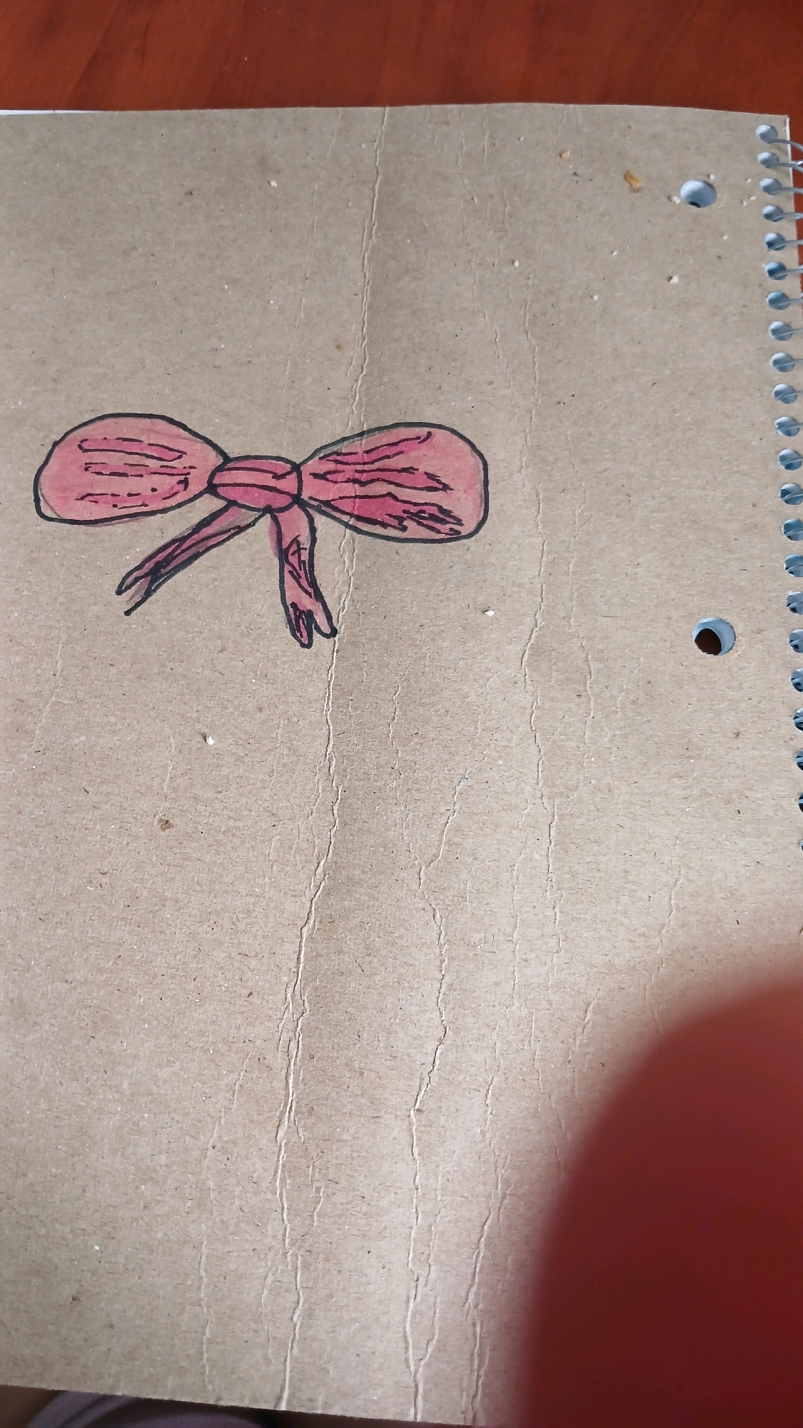 A pink bow is drawn on a brown notebook. The bow is positioned on the left side of the notebook and is colored with pink marker. The notebook is spiral-bound on the right side. The notebook paper has visible lines and some specks of dirt. The background of the image appears to be a wooden table. The location of this notebook in the city of Haivoron, Ukraine is unknown.