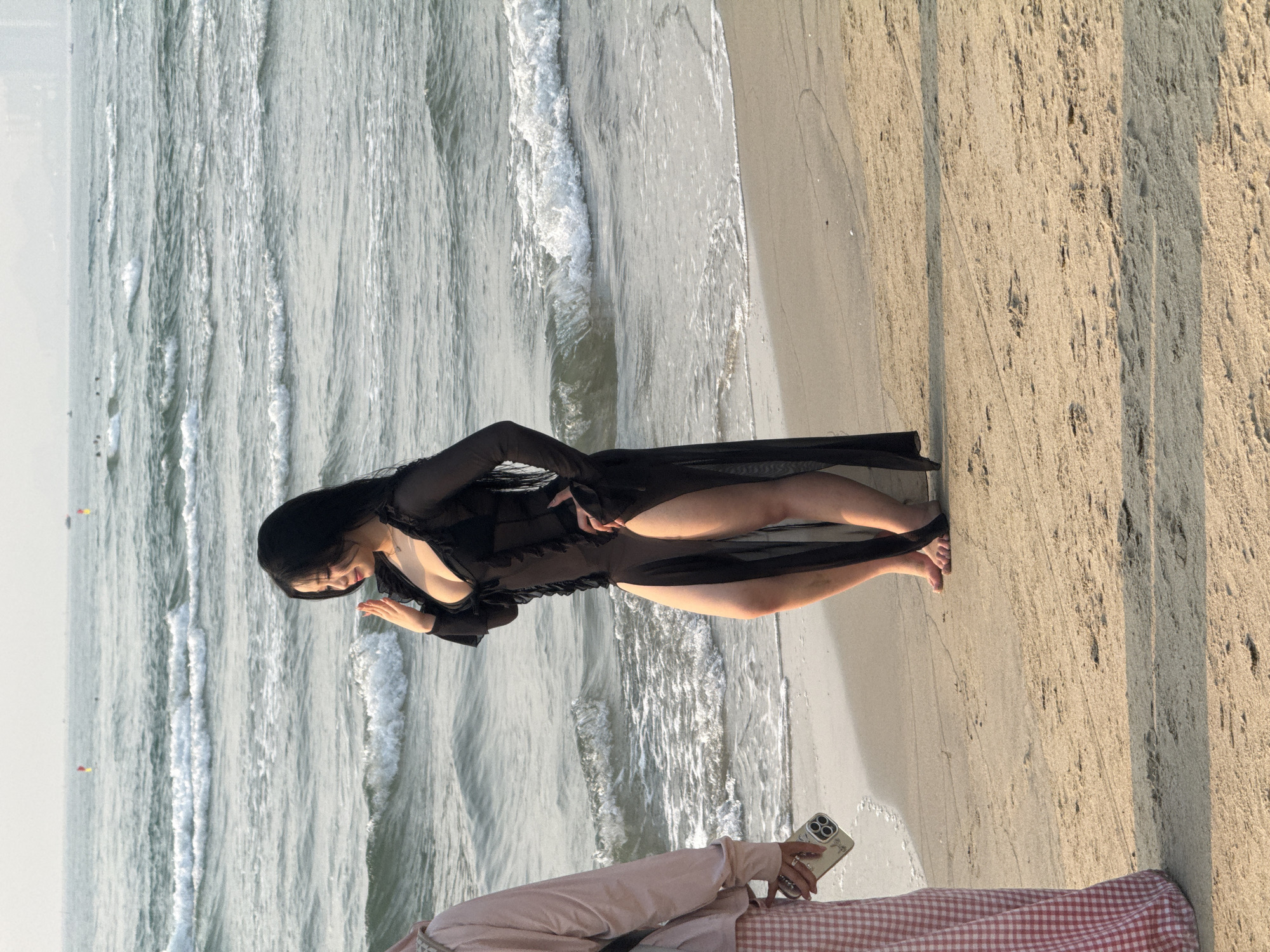 The image shows a woman in a black dress standing at the edge of the ocean, likely in Đà Nẵng, Vietnam, given the location context. She faces the water and appears to be interacting with it, possibly touching or playing with the waves. The ocean has visible waves breaking near the shore. The beach is sandy, and there is another person in the foreground who is partly visible, holding a phone and sitting on a blanket.