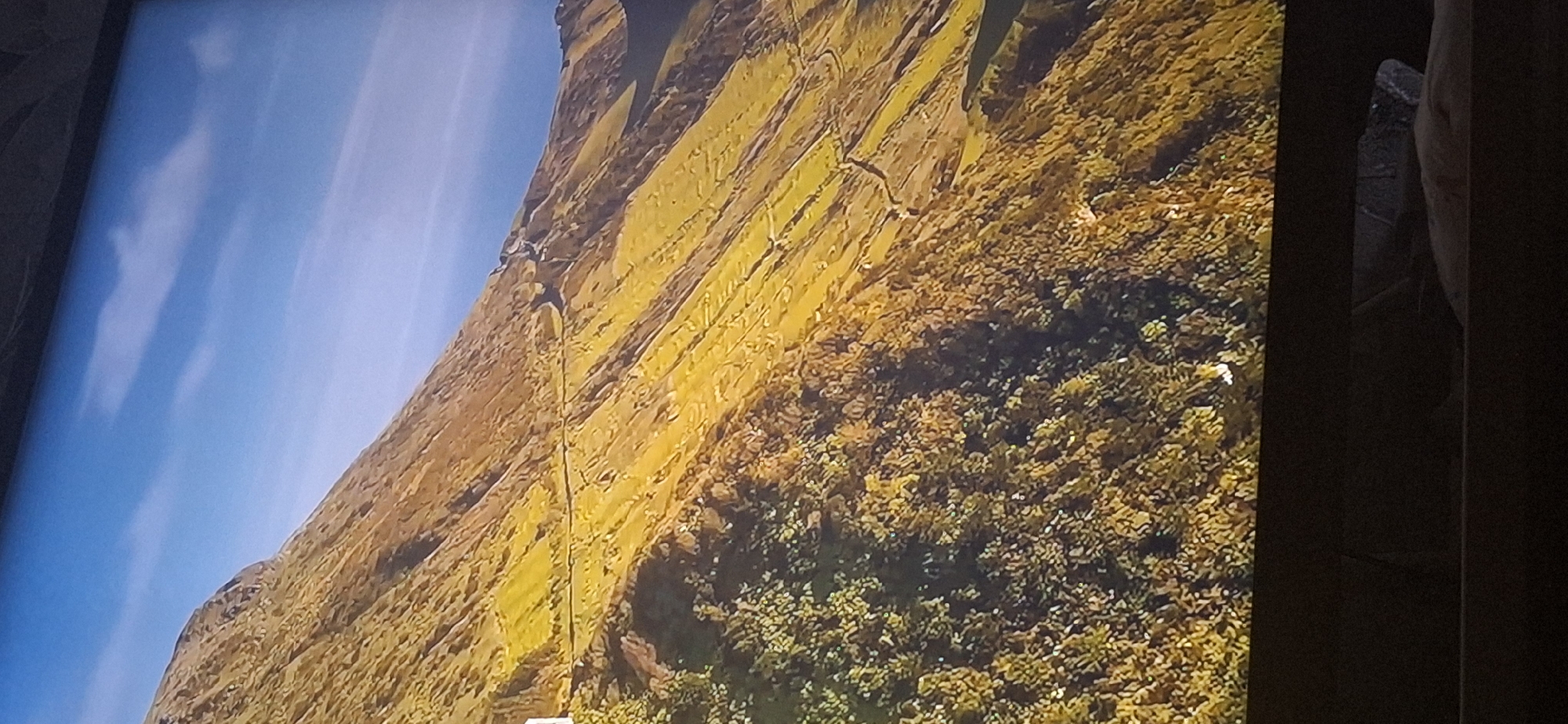 The image shows a steep, grass-covered mountain slope under a bright blue sky with scattered clouds. A winding dirt road or path is visible traversing the side of the mountain. The lower part of the slope is covered in dense green vegetation. The scene appears to be a natural landscape, likely a mountainous region. There is no indication of the city of Bila Tserkva, Ukraine.