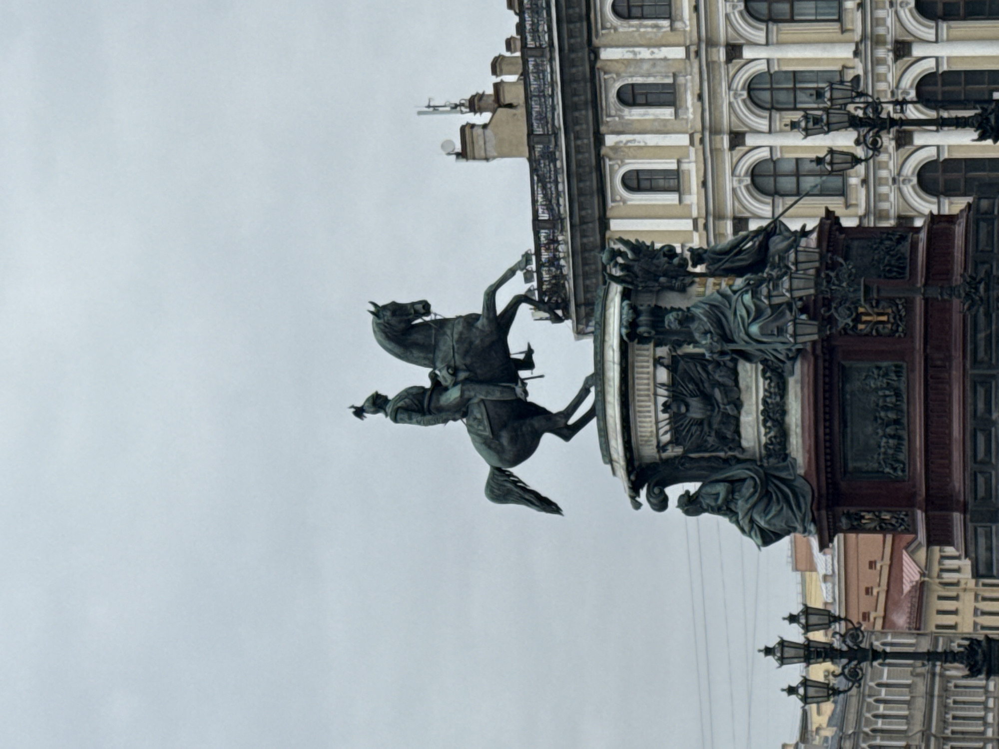 The image, rotated 90 degrees counter-clockwise, depicts a prominent bronze equestrian statue against an overcast sky and a grand classical building. The central subject is an equestrian monument featuring a figure, likely an emperor or military leader, astride a powerfully rearing horse. The rider is depicted with a flowing cloak, his right arm extended and pointing forward, while the horse’s front hooves are dramatically lifted off the ground, conveying motion and authority.
The statue is mounted on a large, elaborate pedestal that is adorned with additional sculptural figures and relief carvings. Several robed, allegorical figures are visible on the lower portions of the pedestal, contributing to the monument's narrative and ornate design. To the right of the statue, a tall, black, multi-lanterned street lamp stands, featuring decorative scrollwork.
Behind the monument, a large, multi-story building dominates the background. Its facade is light-colored (possibly pale yellow or beige) with white architectural details, including prominent cornices and window surrounds. The building features multiple arched windows, set in a rhythmic pattern, indicating a classical or Neoclassical architectural style. The sky is a uniform light grey, suggesting an overcast day. No specific activity is taking place, as the image captures a static monument. There is no visible text. The scene is characteristic of a grand public square or avenue in a historic European city, specifically Saint Petersburg, Russia.