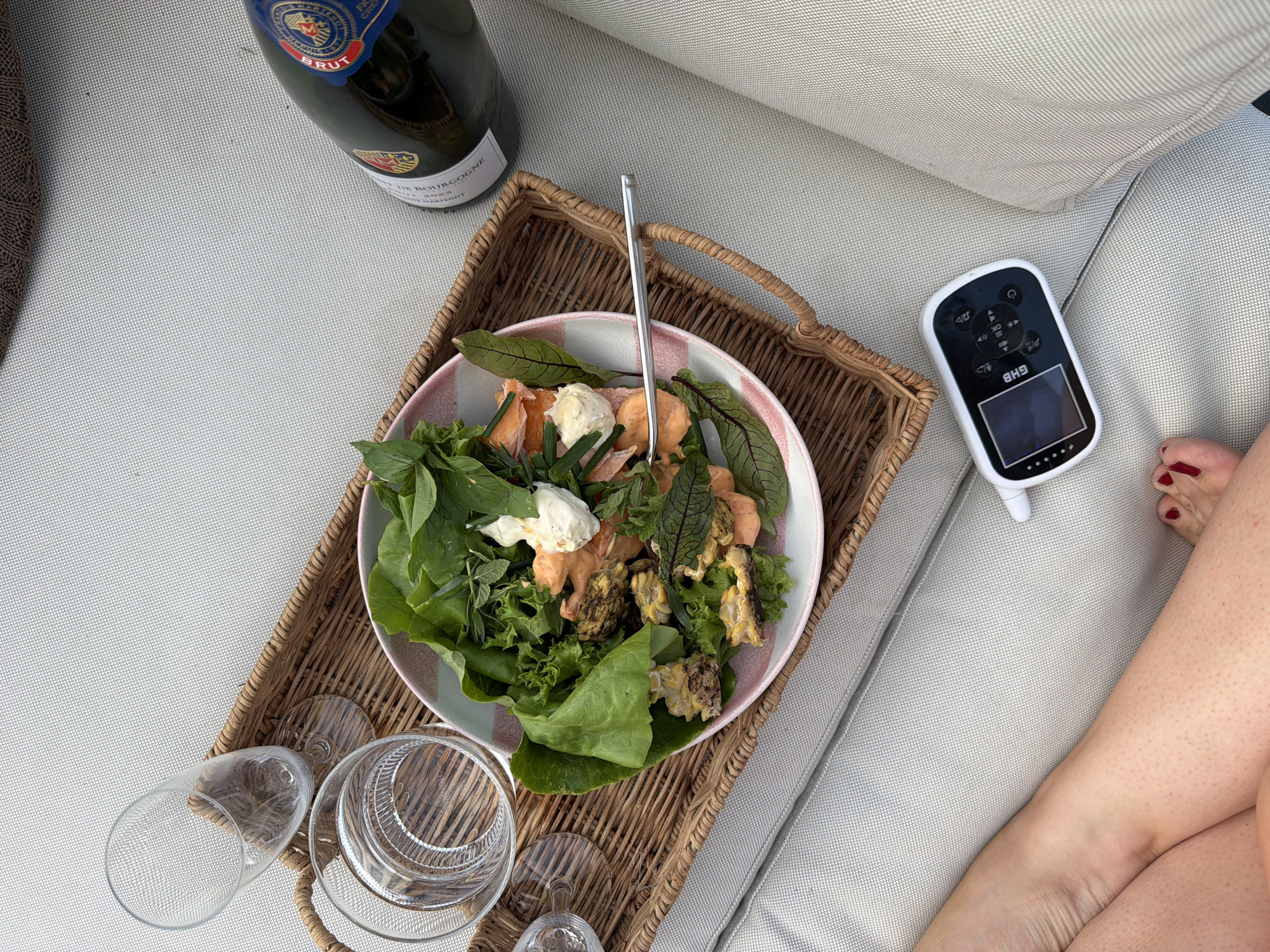 An overhead, high-angle shot depicts a wicker tray on a light-colored outdoor seating area, possibly a patio or deck, on a sunny day. On the tray, a white and pink rimmed bowl holds a salad composed of leafy greens, salmon, and what appears to be corn and other vegetables. A fork rests on the edge of the bowl. Beside the salad are two glasses, one filled with water. A bottle of champagne, with a blue and red label, is also on the tray, positioned to the right. In the lower left corner of the image, a person's legs and feet are visible, suggesting they are seated on the outdoor furniture. To the right of the legs, a small electronic device with a screen and buttons is placed on the seating surface. The overall impression is a relaxed, outdoor meal setting, with no clear indications of the city of Waldgrehweiler, Germany, being present in the image.
