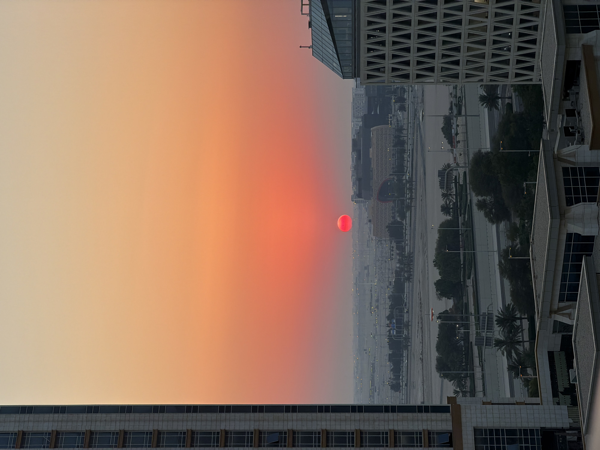 This is a vertical, eye-level shot of a city skyline at sunset. The sky is a gradient of soft orange and peach hues, with a distinct, bright red disc of the sun visible in the middle left of the frame. The city below appears hazy, with numerous buildings of varying heights and architectural styles. The closest building on the right has a white, textured facade and dark-paned windows. The building on the far left has a darker, more modern design with geometric patterns on its exterior and a glass facade.
The scene is urban, suggesting a densely populated area. The haze in the air and the warm tones of the sunset indicate either pollution or a specific atmospheric condition. The overall mood is serene and tranquil, with the sunset lending a peaceful, yet dramatic, atmosphere. The image appears to be taken from a high vantage point, possibly from a hotel room or an upper floor of an office building, looking out over the city. No people are visible, and the focus is on the architecture and the natural phenomenon of the sunset. The time of day is clearly sunset, with the weather being clear enough for a visible sunset, albeit with some atmospheric haze. There are no visible text elements in the image.