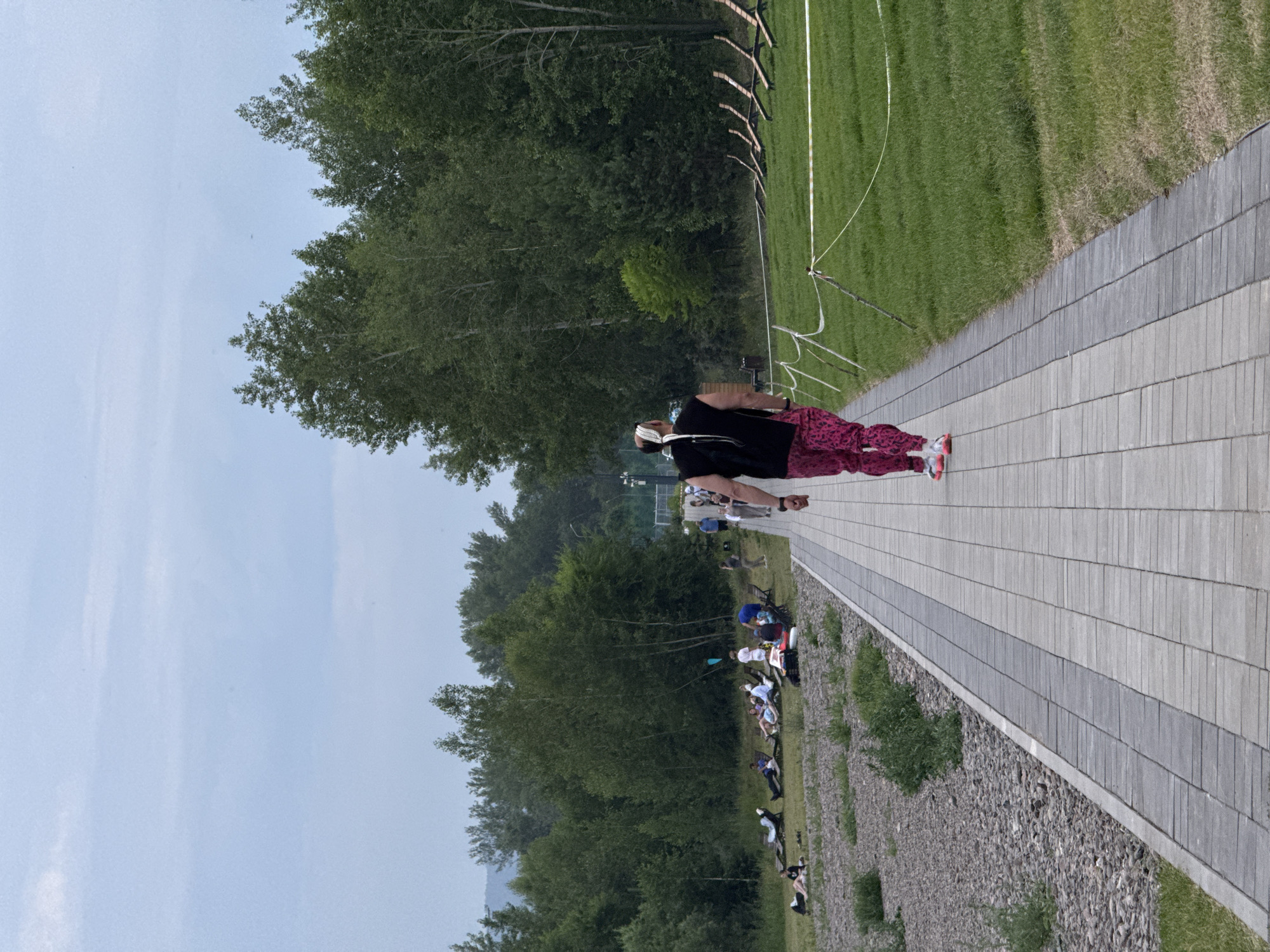 An adult with dark hair and braids, wearing a black sleeveless shirt and pink patterned leggings, walks away from the camera on a paved walkway. They are also wearing pink and white athletic shoes. To the left of the walkway is a gravel area with scattered green vegetation, where several individuals are seated or lying on lounge chairs. Further left, a dense line of trees forms the background. To the right of the walkway is a neatly mowed grassy area, with a white tape barrier supported by wooden stakes. A group of lounge chairs are set up on the grass in the middle distance. In the far distance, more trees are visible, and a misty mountain range can be seen. The sky is overcast and light blue. The scene appears to be a park or recreational area, possibly near a body of water given the presence of lounge chairs in a public area, and the general landscape suggests an outdoor public space. This park is located in the city of Krasnoyarsk, Russia.