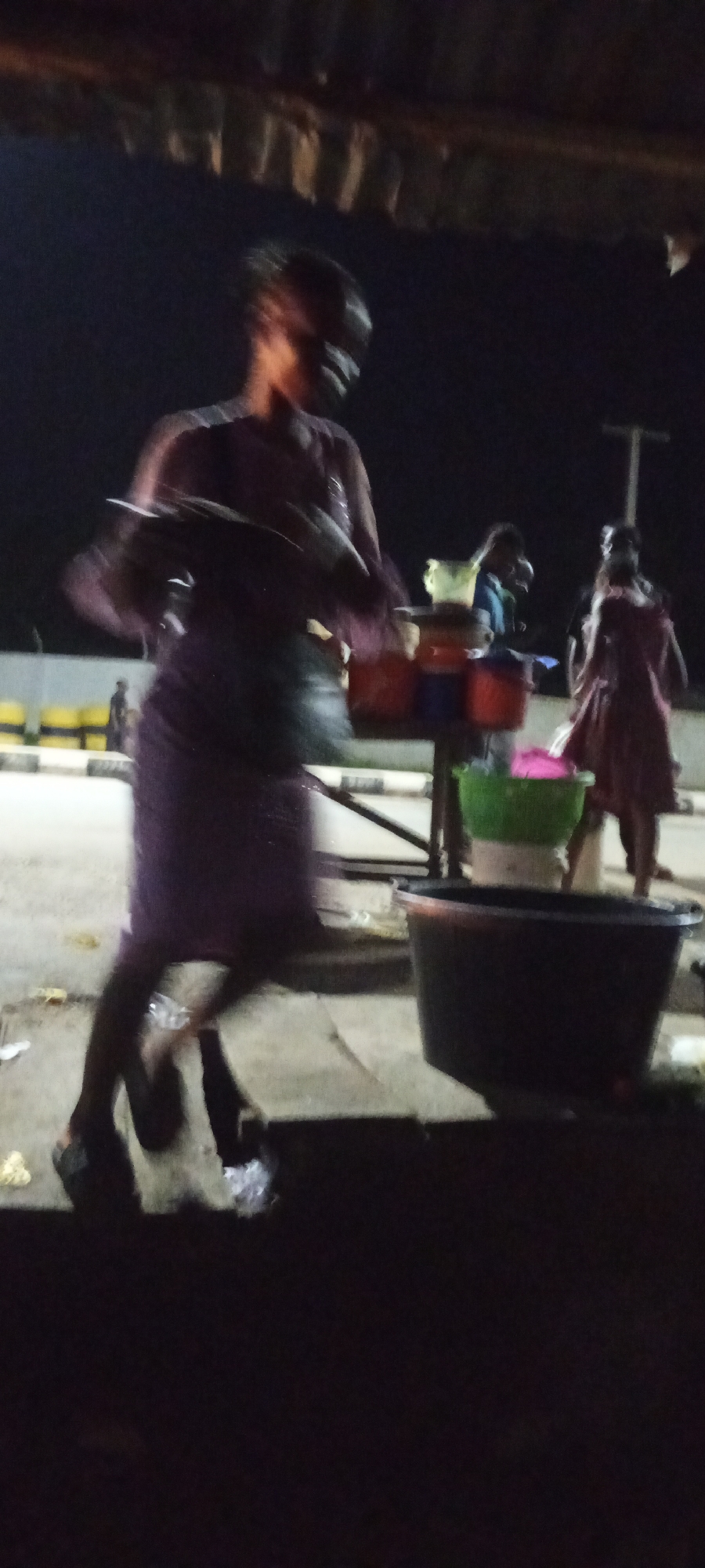 An outdoor night scene in Adankolo, Nigeria shows a woman in a purple dress with a dark bag on her shoulder, her face is blurred. She is wearing black shoes. She is walking past several large plastic basins, one of which is black and appears to be a large tub. Behind her, other individuals are visible in the background, some of whom are also blurred. The setting appears to be a street market or roadside stall, indicated by the presence of the basins and the covered structure above. The ground is uneven and appears to be paved, with some scattered debris. The lighting is dim, suggesting artificial light sources are present.