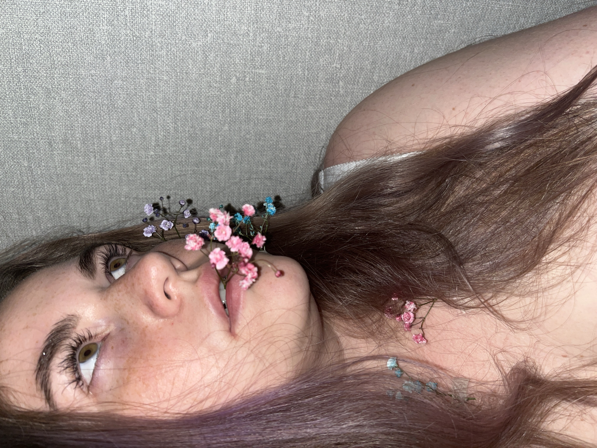 A close-up image features a fair-skinned person lying on what appears to be a light gray or beige textured surface, possibly a couch or bed. The person's face is turned slightly upward, with their gaze directed towards the upper left of the frame. Their hair, which has brown and purple tones, is spread around their head and shoulders. Small pink, blue, and purple flowers are positioned near their mouth and scattered in their hair. The image focuses on the person's face, capturing details such as freckles, eyelashes, and the shape of their lips and nose. There is no information about a specific location in Chaikovsky, Russia, or further context in the image.