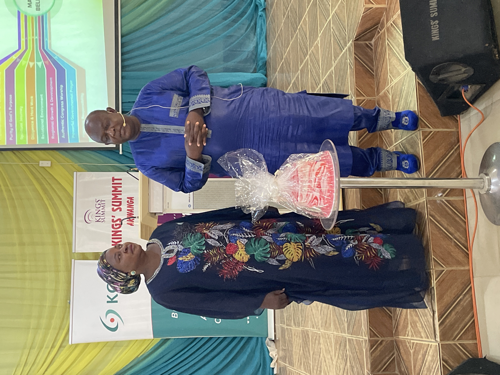 A photograph shows two people, a man and a woman, in front of a banner for the Kings' Summit in Akwanga. The woman is on the left, wearing a dark blue dress with floral embroidery and a patterned head covering. Next to her is a stand holding a gift wrapped in clear plastic. The man, standing slightly behind her on the right, wears a matching blue traditional robe and cap. He has a microphone clipped to his shirt.
Behind them is a screen displaying a presentation slide titled "Purity of God's Purpose" with bullet points. The background includes a curtain with blue and yellow fabric draping and a tiled wall and floor. A large black speaker is in the upper right corner.