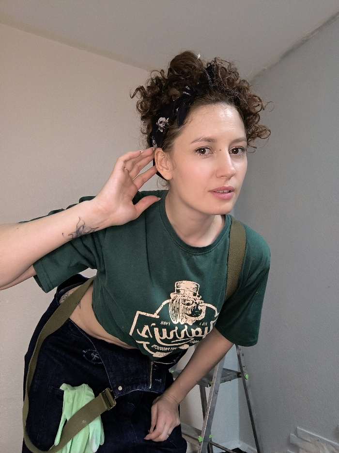 A female subject is captured in an indoor, waist-up portrait. She has light brown skin, curly dark brown hair styled upward and secured with a black bandana featuring white skull patterns. Her eyes are light brown, and she has a small blemish on her left cheek. She wears a dark green, short-sleeved crop top displaying a light-colored, reversed graphic of a dog in a top hat with text including "SINNEN 1880 FINEST PRODUCTS." She is also wearing dark blue denim overalls, with a khaki-colored strap crossing her torso diagonally. A light green glove is tucked into the left side of her overalls. Her left forearm bears a small tattoo. A silver ring is visible on her left ring finger. She holds her right hand with fingers near her right ear, facing the camera with a direct gaze and neutral expression. The background features plain walls, with sections appearing light beige and light gray. A metallic step ladder is partially visible on the lower right side of the frame. The setting suggests an interior space, possibly a residence or workshop undergoing renovation, located within Saint Petersburg, Russia; the specific address or detailed locale is not discernible.