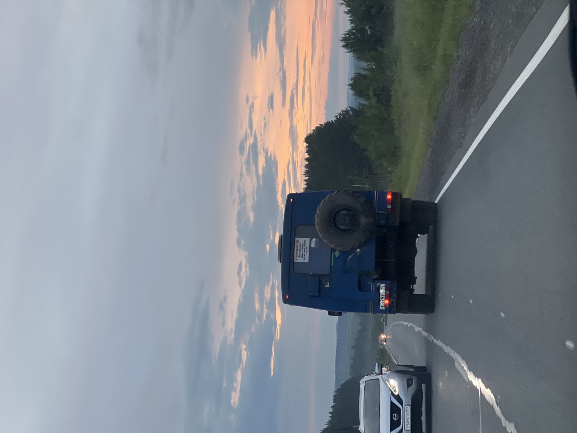 The image captures a view from a vehicle traveling on a paved road during dusk. A blue van with a spare tire mounted on its rear is in the foreground, to the right of the lane. A white SUV is in the left lane, ahead of the viewer's vehicle. The sky is filled with a gradient of colors, transitioning from light blue to soft oranges and pinks as the sun sets. Clouds are scattered across the sky. The road is bordered by trees and a grassy embankment on the right. The overall scene suggests a journey through a rural or semi-rural area during twilight. There is no indication of the specific location in Doksha, Russia.