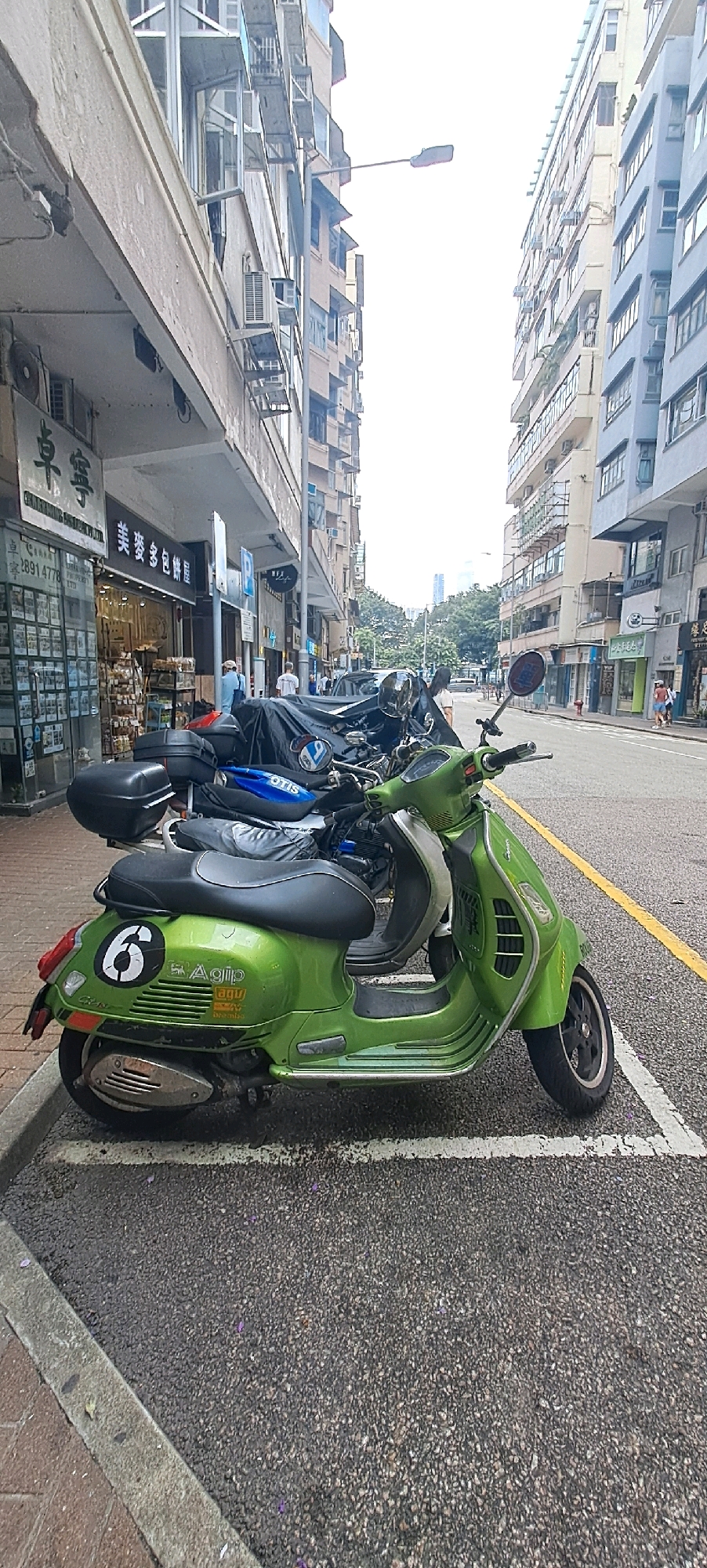 The image shows a street scene in Hong Kong, China. In the foreground, a bright green Vespa scooter is parked in a designated parking spot. The scooter has a black seat and a "6" sticker on its rear panel. Behind the Vespa, other motorcycles are parked, some covered with tarps. To the left of the motorcycles are storefronts with various signs in Chinese characters and some English text. One sign reads "美麥多包餅屋" (Mei Mak Duo Bao Bing Wu), indicating a bakery. Further down the street, buildings line both sides, characteristic of Hong Kong's dense urban environment. People are visible walking on the sidewalks and crossing the street in the distance. The sky is overcast, and the street appears wet, suggesting recent rain. The scene depicts a typical day in a busy commercial area of Hong Kong.