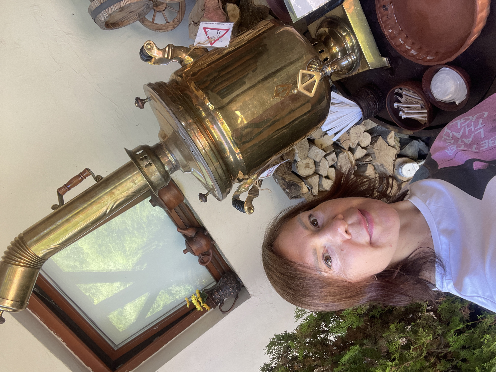 A woman with shoulder-length brown hair takes a selfie in front of a large, ornate brass samovar. The samovar is positioned on a table with various items, including stacked firewood, a small barrel with wheels, a miniature teapot, a vase with yellow flowers, and bowls containing sugar packets and possibly lids. A caution sign with a graphic of a steaming object is attached to the samovar. The background includes a window with a view of green foliage and a textured wall. The woman is wearing a white t-shirt with a pink and black design. The scene appears to be outdoors or in an open-air setting, possibly a cafe or market in Estosadok, Russia, given the cultural context of the samovar.