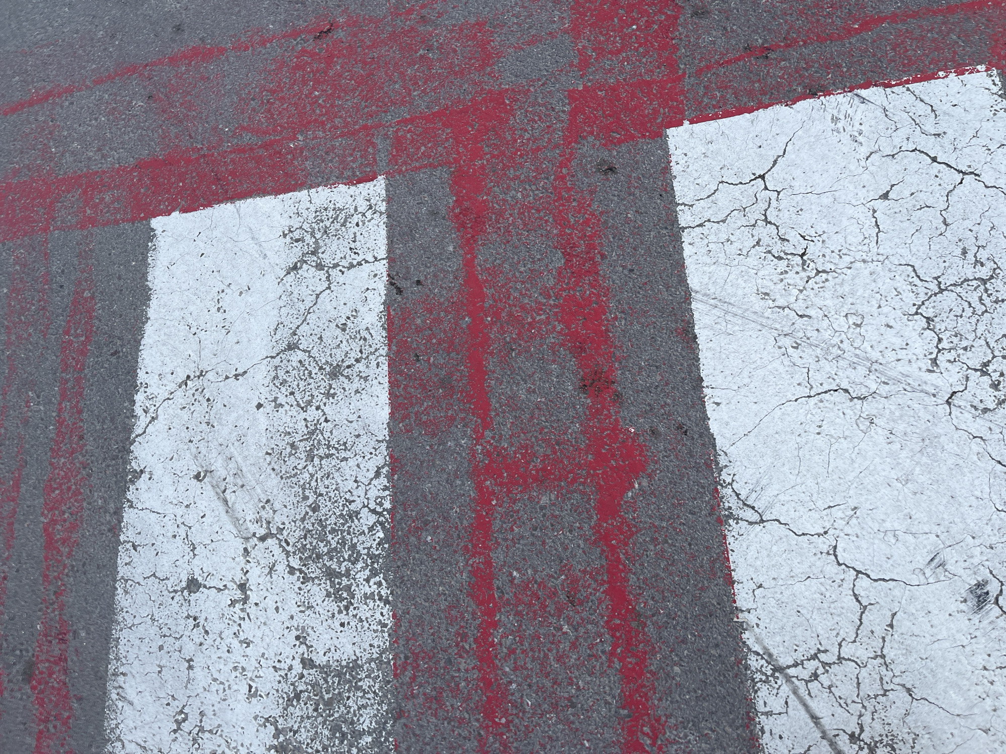 The image shows a section of asphalt pavement in what appears to be a crosswalk or pedestrian crossing. The asphalt is gray and shows signs of wear and tear. The crosswalk is marked with alternating white and gray stripes. The white stripes are cracked and worn. Red paint has been applied over parts of the crosswalk, obscuring some of the white and gray stripes. The red paint appears faded and worn, with vertical streaks. The scene is devoid of people. Without additional information, it's impossible to confirm the specific location within Budva, Montenegro, but the markings suggest an urban environment with pedestrian traffic.