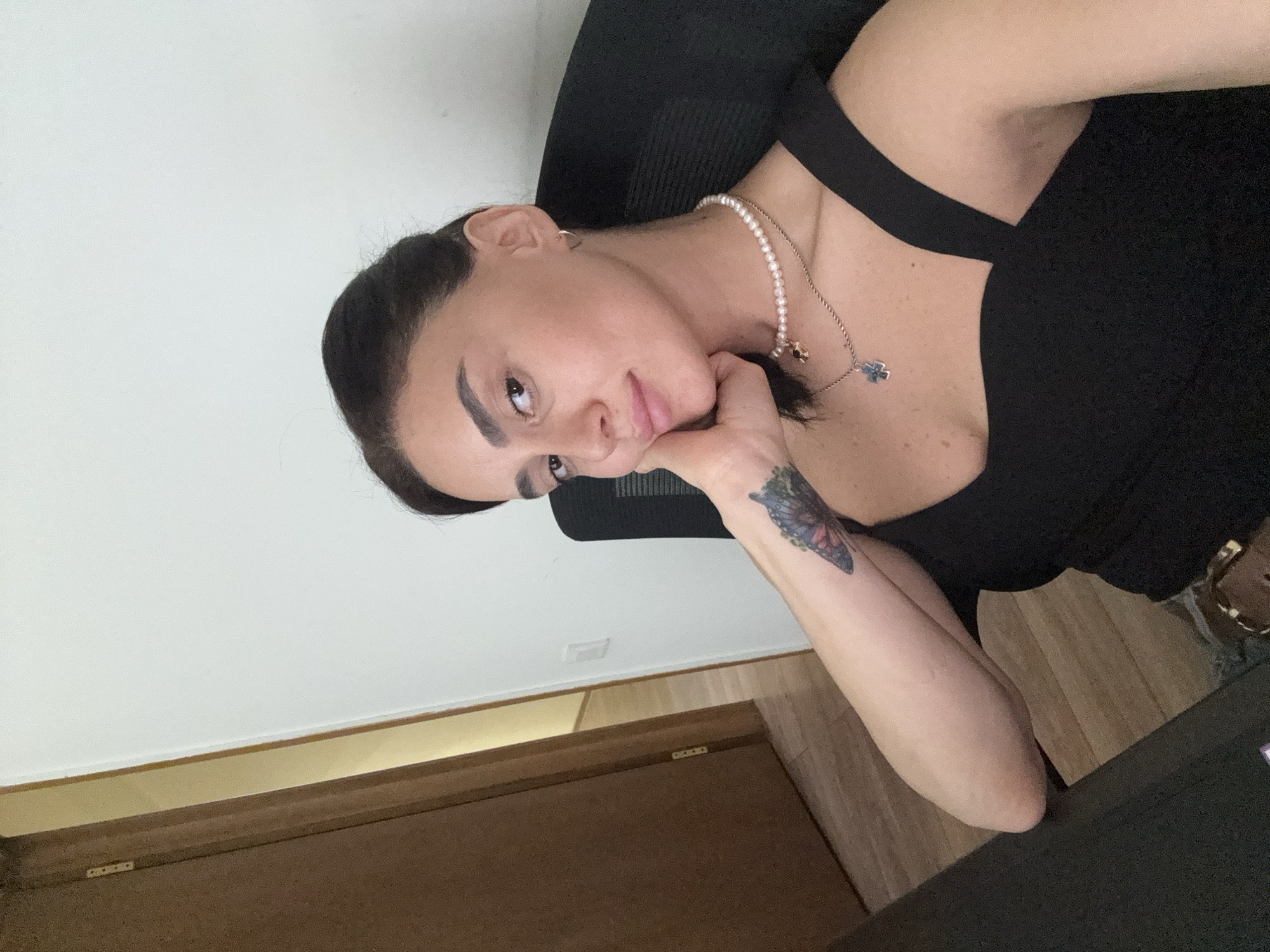 A woman with dark hair and makeup is sitting at a desk, looking at the camera. She has a butterfly tattoo on her left forearm and is wearing a black top with a pearl necklace and a pendant. The background shows a doorway and a white wall.