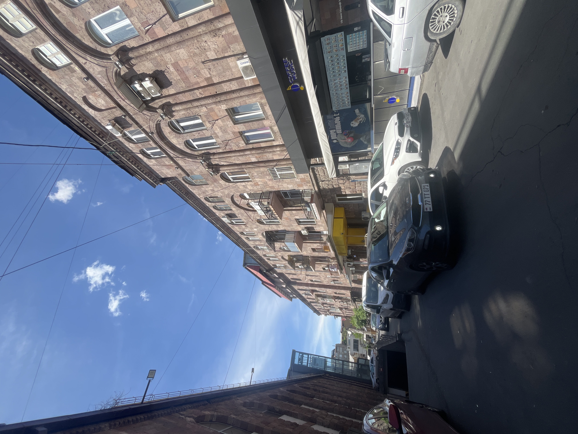 The image shows a street in Yerevan, Armenia, with buildings on either side. The buildings are made of brick and have multiple windows and balconies. There are several cars parked along the street, including a black Kia with the license plate "77 TT 071". There is also a small building with glass and metal construction along the street. The sky is blue with some clouds. There's a coffee shop with the name "Coffee Mood" on one of the buildings.