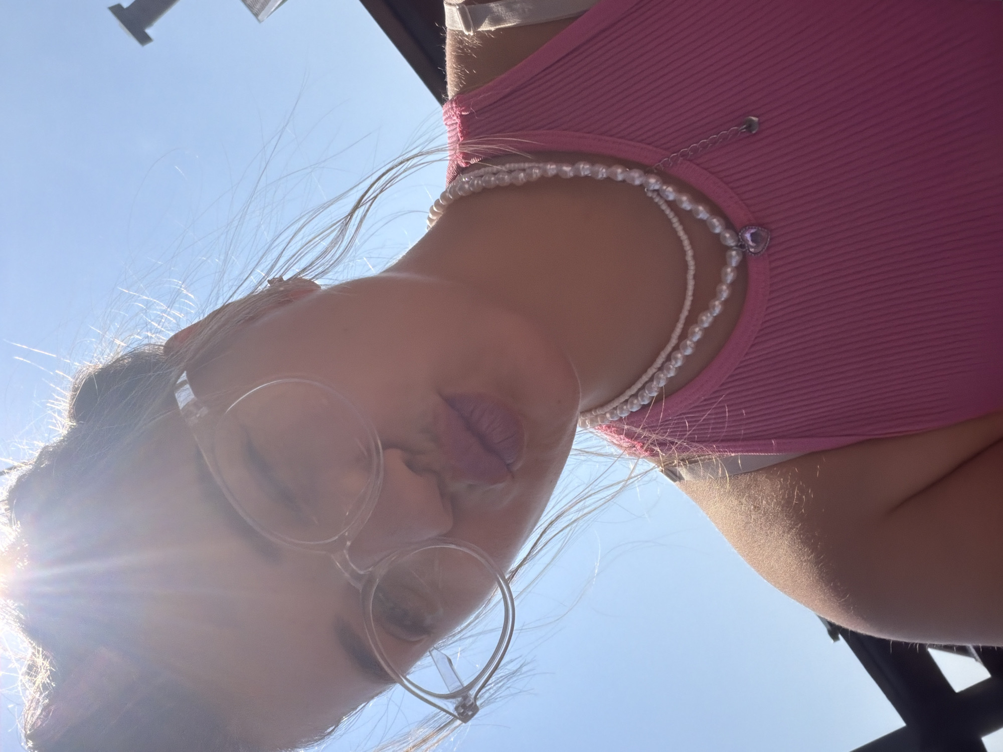 A woman, viewed from a low angle looking upward, is wearing round glasses and a pearl necklace with a heart pendant. She has fair skin and wears a pink ribbed tank top. The background is a clear blue sky with a bright sun glare illuminating the lower left corner. The location appears to be outdoors on a sunny day, based on the sky conditions. There is some dark structure on the bottom right corner. There is no other evidence that suggests this picture was taken in Большая Шильна, Russia.