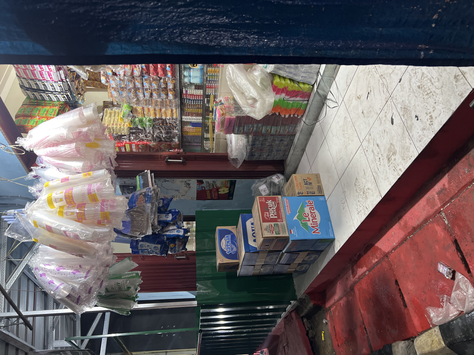 The image shows the exterior of a small convenience store in Citeureup, Indonesia. The storefront is open, revealing shelves stocked with various goods. In the foreground, there are stacks of bottled water, including "Le Minerale" and "AQUA," as well as boxes of tea. Plastic cups are displayed hanging from above the entrance. Bags of what appear to be snacks or other packaged goods are also visible on shelves inside the store. The ground outside the store is tiled and appears somewhat wet, with a red-painted drainage area. The overall scene depicts a typical street-level retail establishment in Indonesia, likely operating in the evening given the lighting.
