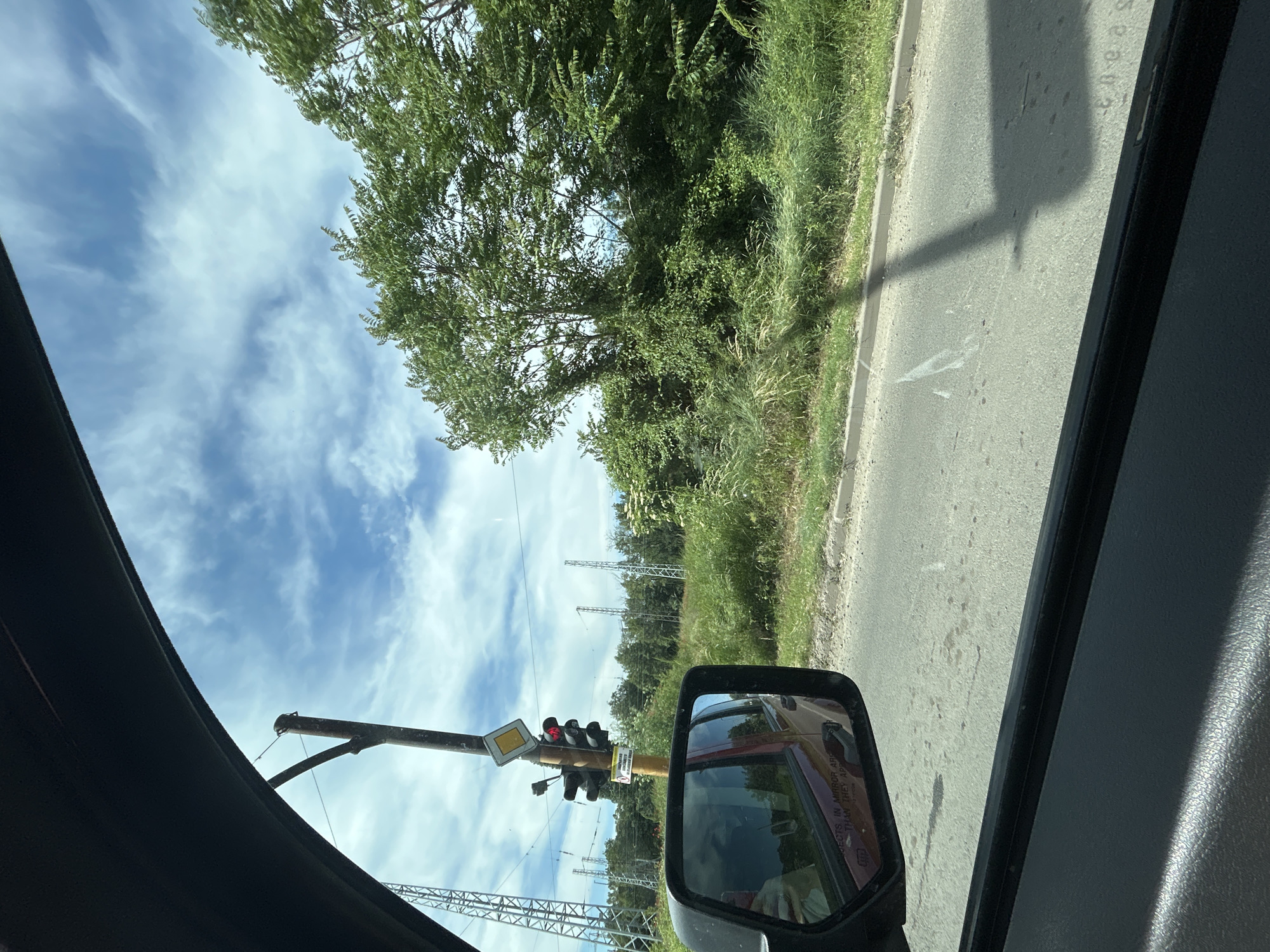 The image is a view from inside a vehicle, likely a car, showing a road and roadside vegetation, along with electrical transmission towers in the distance. A side mirror is visible in the foreground. A traffic light with red lights is located in the background. The location cannot be identified as Sofia, Bulgaria, from the information in the picture. The time of day is daytime. Weather is bright.