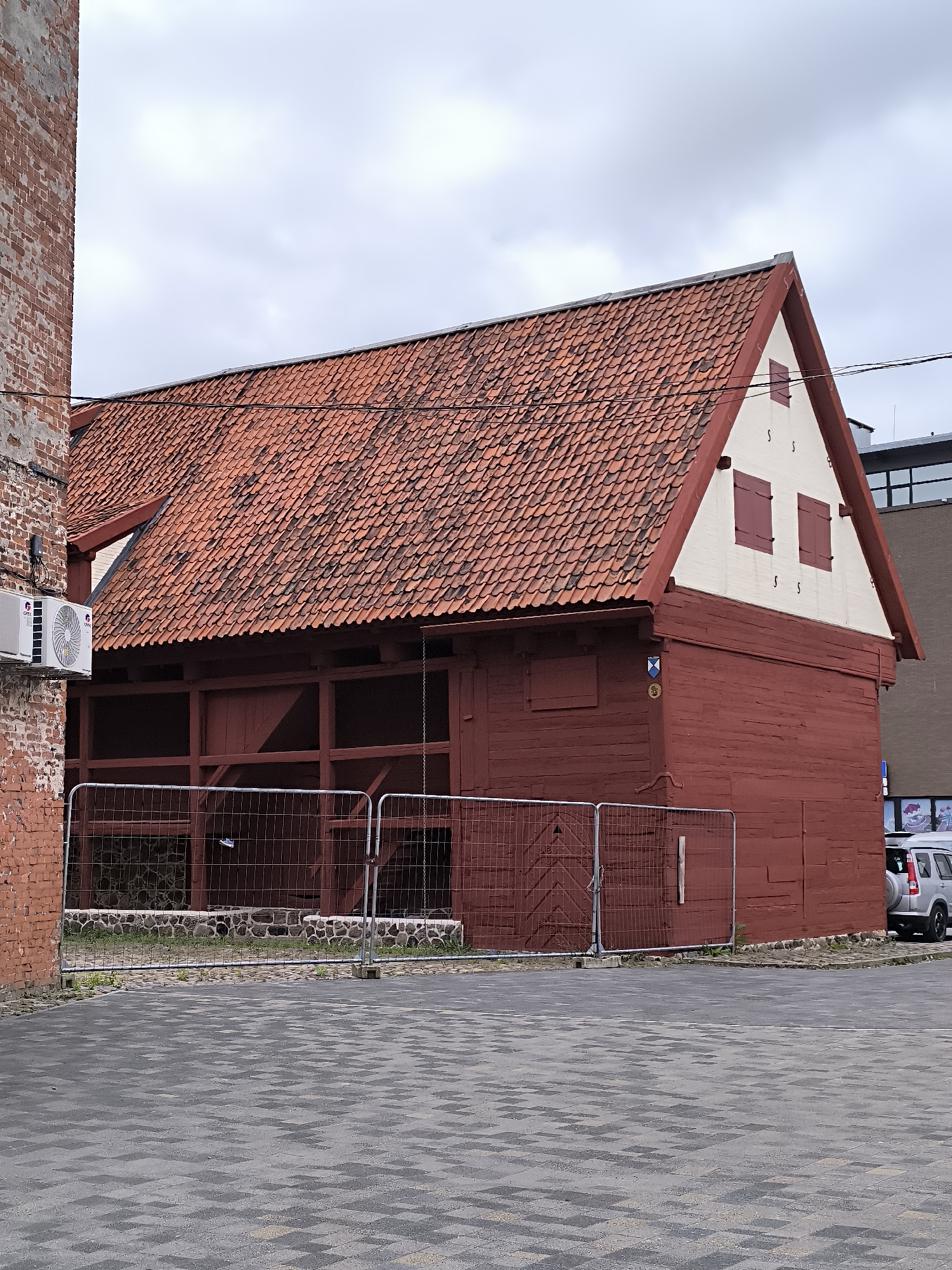 The image captures a two-story, deep red wooden building with a red-tiled gabled roof, situated in the city of Liepaja, Latvia. The building appears to be an old barn or warehouse, with exposed wooden beams visible on the lower level through a fence. The upper section of the building is painted cream with two red-shuttered windows and decorative 'S' shapes. A weathered brick wall, part of another structure, stands to the left of the wooden building. In front of the wooden building, a temporary metal fence encloses the area. A silver SUV is parked to the right of the building. The ground is paved with dark grey interlocking bricks. The sky is overcast.