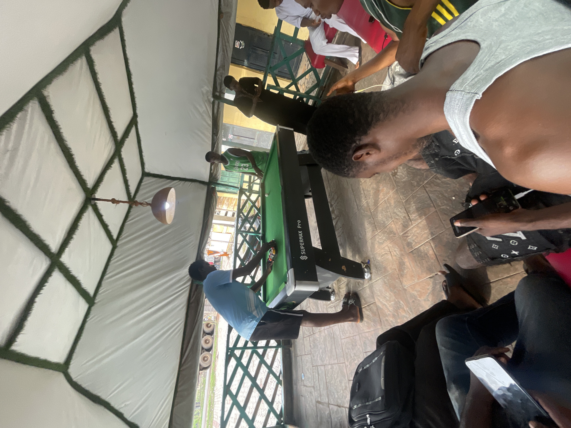 Location: Outdoor space in Lokoja, Nigeria.
Scene: Several individuals are gathered around a pool table labeled "SUPERMAX PRO." One person wearing a blue shirt is setting up the pool balls on the table. Other individuals are standing or sitting around the table, some using their phones.
Objects: Pool table, pool balls, phones, chairs, and a black backpack.
Environment: The area is covered by a tent-like structure with green framing.