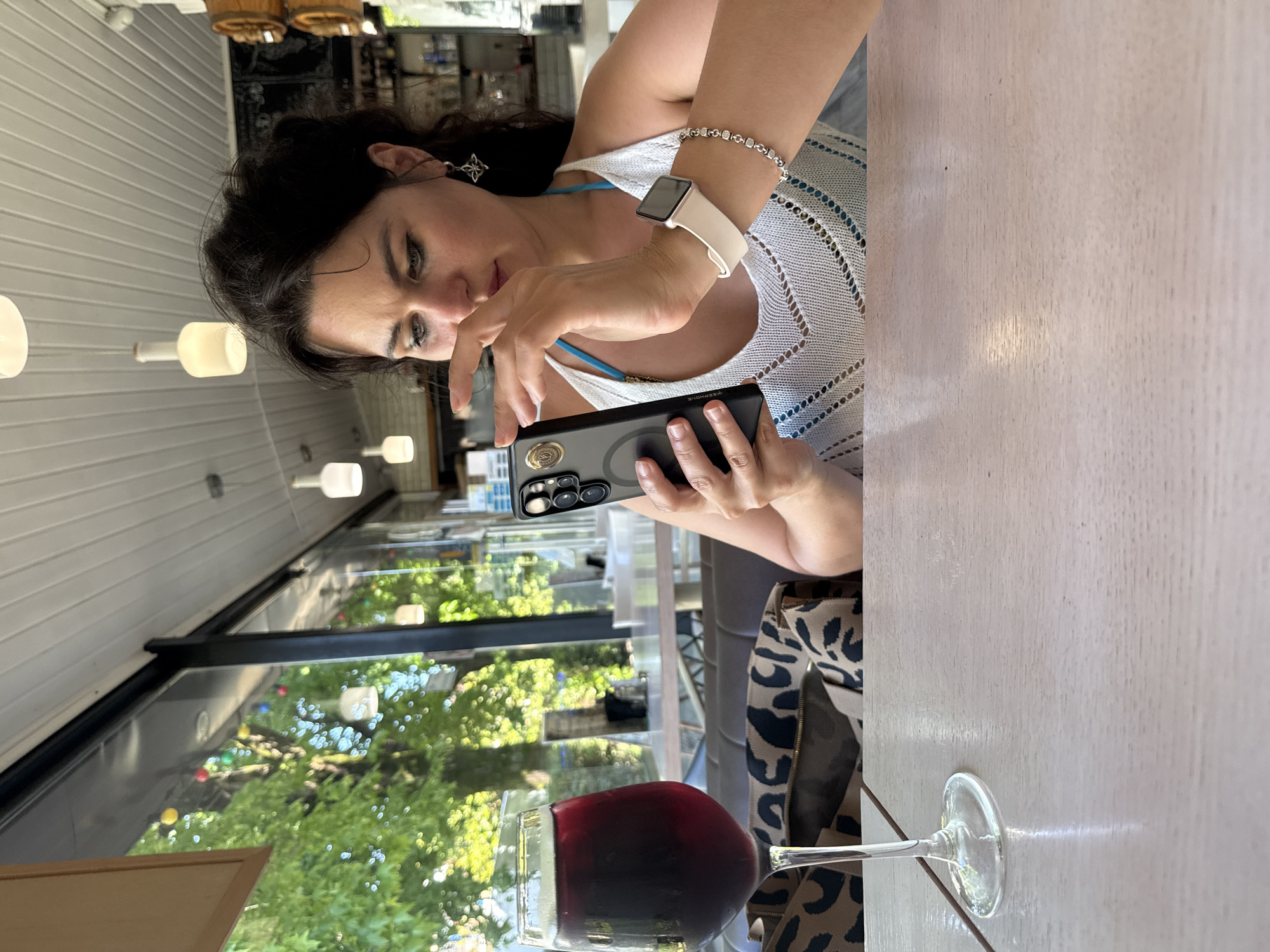 A woman with dark hair sits at a table in a restaurant with a large window overlooking trees and streetlights. She is holding a smartphone and appears to be looking at it. A glass of dark red wine sits on the table in front of her, next to a bag with a leopard print. The restaurant interior is visible behind her, with white ceiling panels and hanging lights. This scene is located in Sochi, Russia.