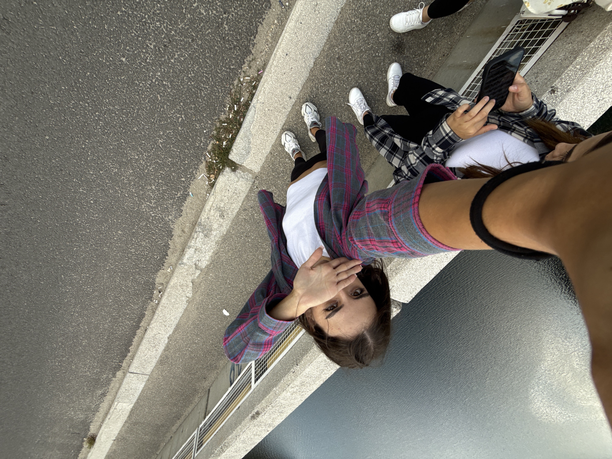 Two young women are standing on the edge of a dam overlooking a body of water. One woman, in the foreground, is taking a selfie with her arm extended, holding a phone. She is wearing a white tank top and a pink and gray plaid shirt. The other woman, slightly behind her, is wearing a black and white plaid shirt, a white tank top, and black pants. She is looking down at her phone, which she is holding in her left hand. Both women are wearing white sneakers. The dam appears to be made of concrete and has a metal railing along the edge. The ground is paved with asphalt. The scene is set in Arefu, Romania.