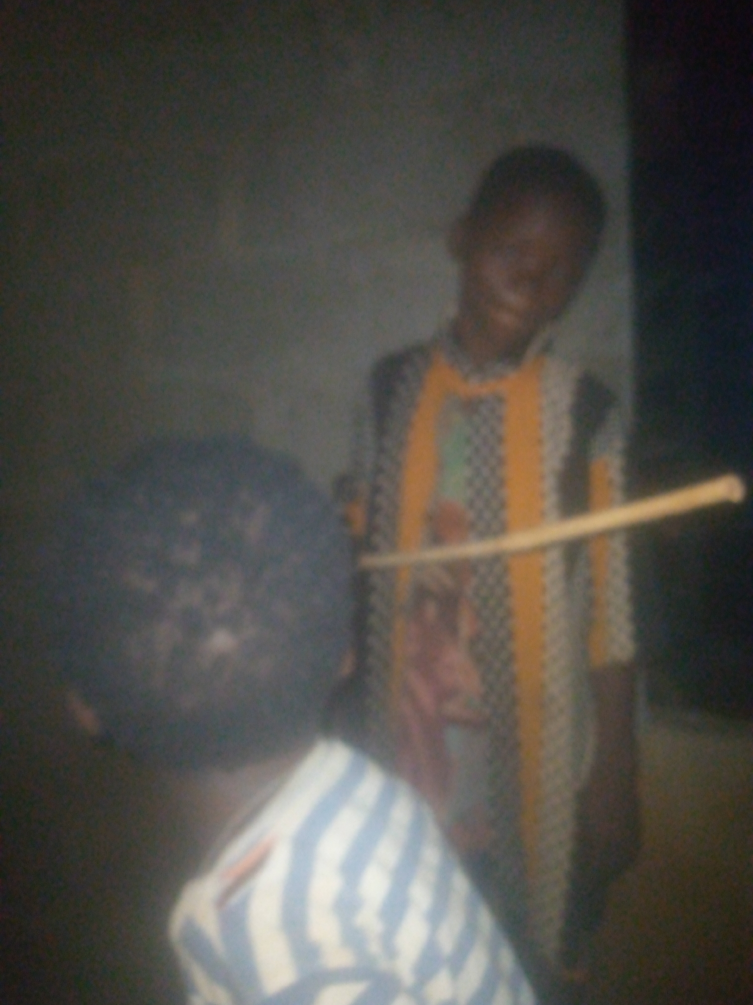The image shows two children indoors. The child in the foreground is wearing a blue and white striped shirt, with their back to the camera. Behind this child, another child is holding a thin, light-colored stick, possibly pointed towards the first child. The second child is wearing a patterned shirt with yellow and grey stripes. The environment is dimly lit, with walls visible in the background. The location is unspecified.