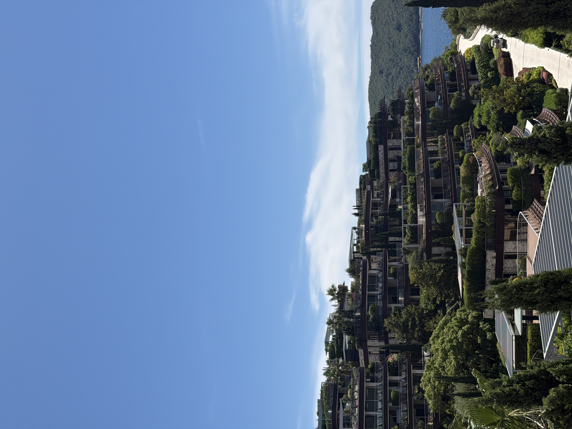 The image depicts a modern residential complex with terraced buildings built into a hillside overlooking the Adriatic Sea in Budva, Montenegro. The architecture features a contemporary design with extensive use of glass, dark-colored facades, and lush greenery cascading down the multiple levels. Each tier appears to have balconies and terraces adorned with potted plants, trees, and flowering shrubs, integrating the structures harmoniously with the natural landscape.
The buildings are situated on a steep incline, with pathways and roads winding through the property. A small white vehicle is visible on one of the roads. The complex is surrounded by dense vegetation, including cypress trees, palm trees, and various bushes, suggesting a well-maintained, landscaped environment.
In the background, the deep blue of the Adriatic Sea stretches towards a distant, verdant hillside covered in trees. The sky above is a clear, bright blue with a few wispy white clouds. The overall impression is one of luxurious, modern living integrated with a beautiful coastal environment. The specific location within Budva is likely a prime spot with panoramic sea views, typical of exclusive residential developments in the area.