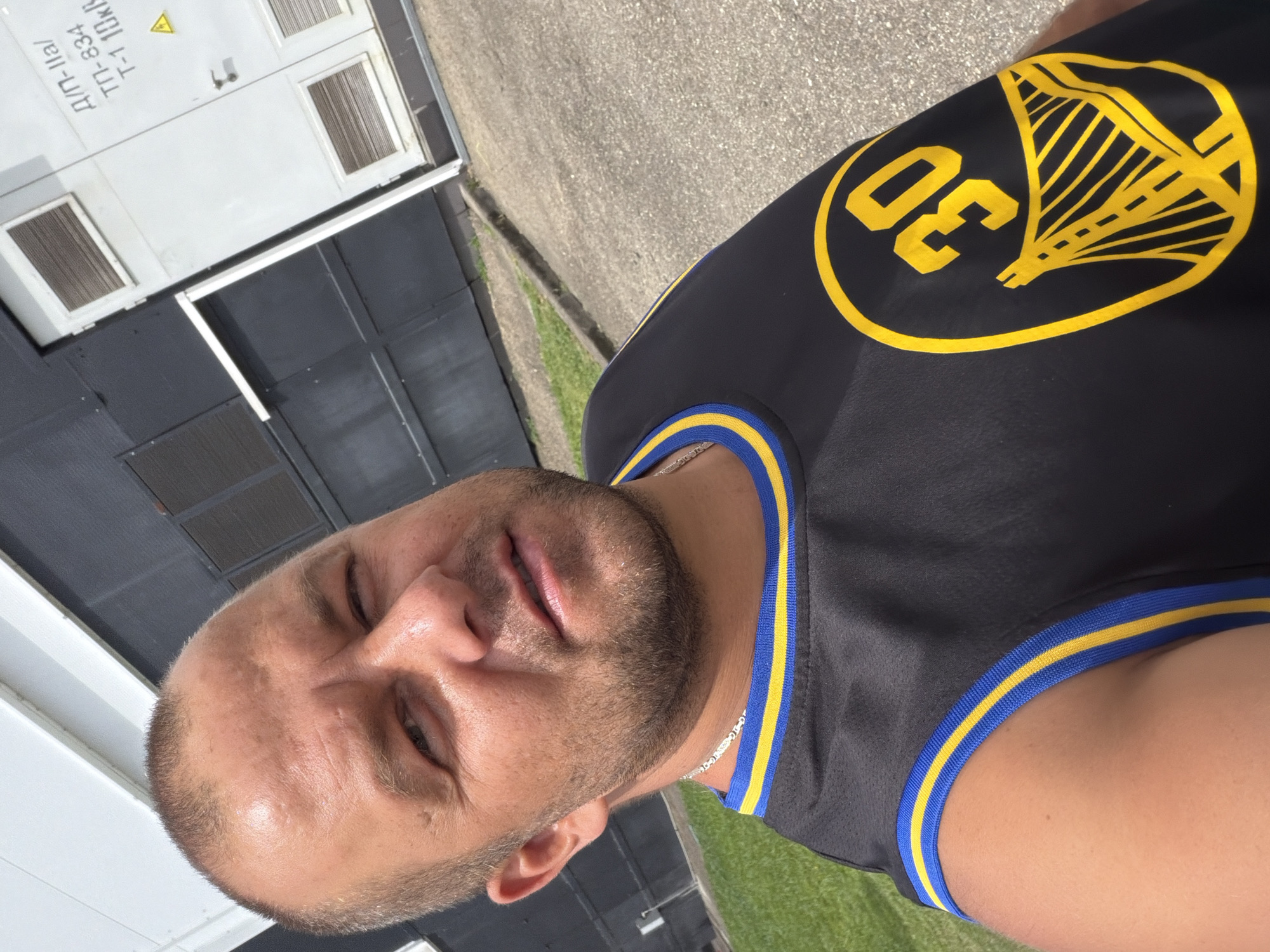 A man is taking a selfie outdoors. He is wearing a black basketball jersey with yellow and blue trim, and a yellow graphic with the number "30" and a bridge design on it. He has short brown hair and stubble. He is wearing a silver chain. The background consists of a building with a dark gray and white facade, and a paved area with some grass and a curb. The building has ventilation grates and markings in Cyrillic script. The image appears to have been taken during the day, with bright sunlight. There is no visible indication of the specific city of Sochi, Russia.