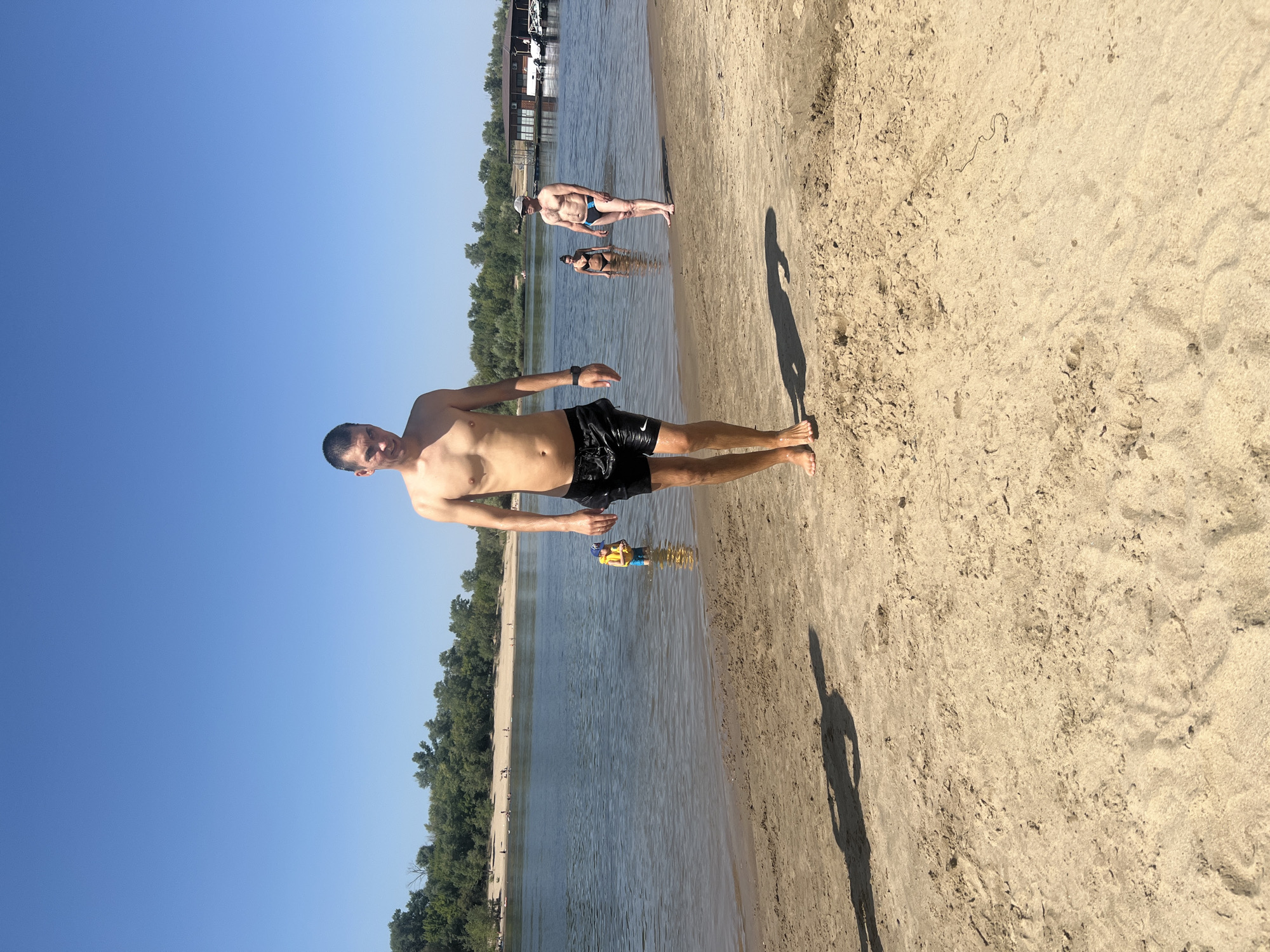 This is a vertically oriented, close-up photograph taken on a sandy beach next to a body of water. The primary subject is a man in black swim trunks standing on the sand, facing the camera. He appears to be in good health and has a tan. His left arm is raised slightly, and he is looking directly at the camera with a neutral expression.
In the background, the water is calm and reflects the clear blue sky. There are other people visible in the water and on the far bank of the river. A man in swim trunks and a woman in a bikini are standing in the water. Further in the distance, along the opposite shore, there is a line of lush green trees. There are also some structures on the far bank, possibly buildings or boats.
A small child or person wearing a yellow floatation device is in the water closer to the foreground. The sand appears dry and textured, with some footprints visible. The sun is casting long shadows, suggesting it is either early morning or late afternoon. The overall mood is relaxed and summery, indicative of a day at the beach. There is no visible text in the image. The scene is consistent with a typical summer day at a river beach in Kilyakovka, Russia.