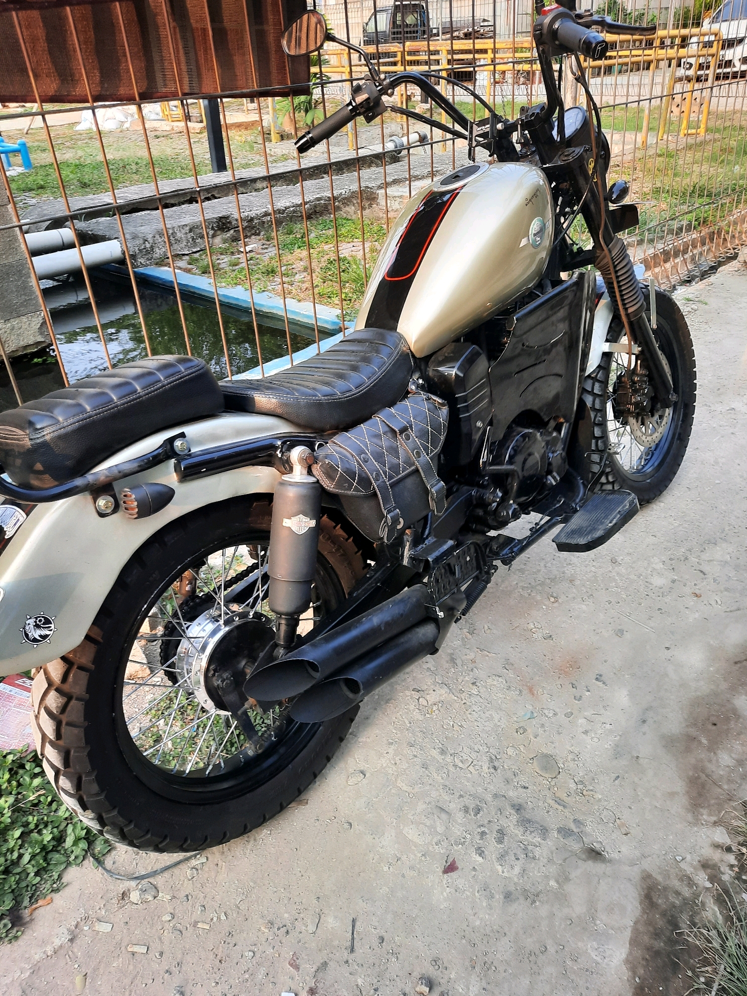 A custom motorcycle, likely a cruiser or bobber style, is parked outdoors on a concrete surface. The motorcycle features a light gold fuel tank with black and red racing stripes, black leather seats, and a black leather saddlebag with diamond stitching. It has a dual exhaust system and knobby tires, suggesting it may be modified for off-road use or aesthetic purposes. The background shows a metal fence, some vegetation, and a shallow body of water with pipes running into it. The scene appears to be in a semi-urban or industrial area, possibly within the city of East Jakarta, Indonesia, given the typical setting for such motorcycles and surrounding environment. There are no people visible in the image.
