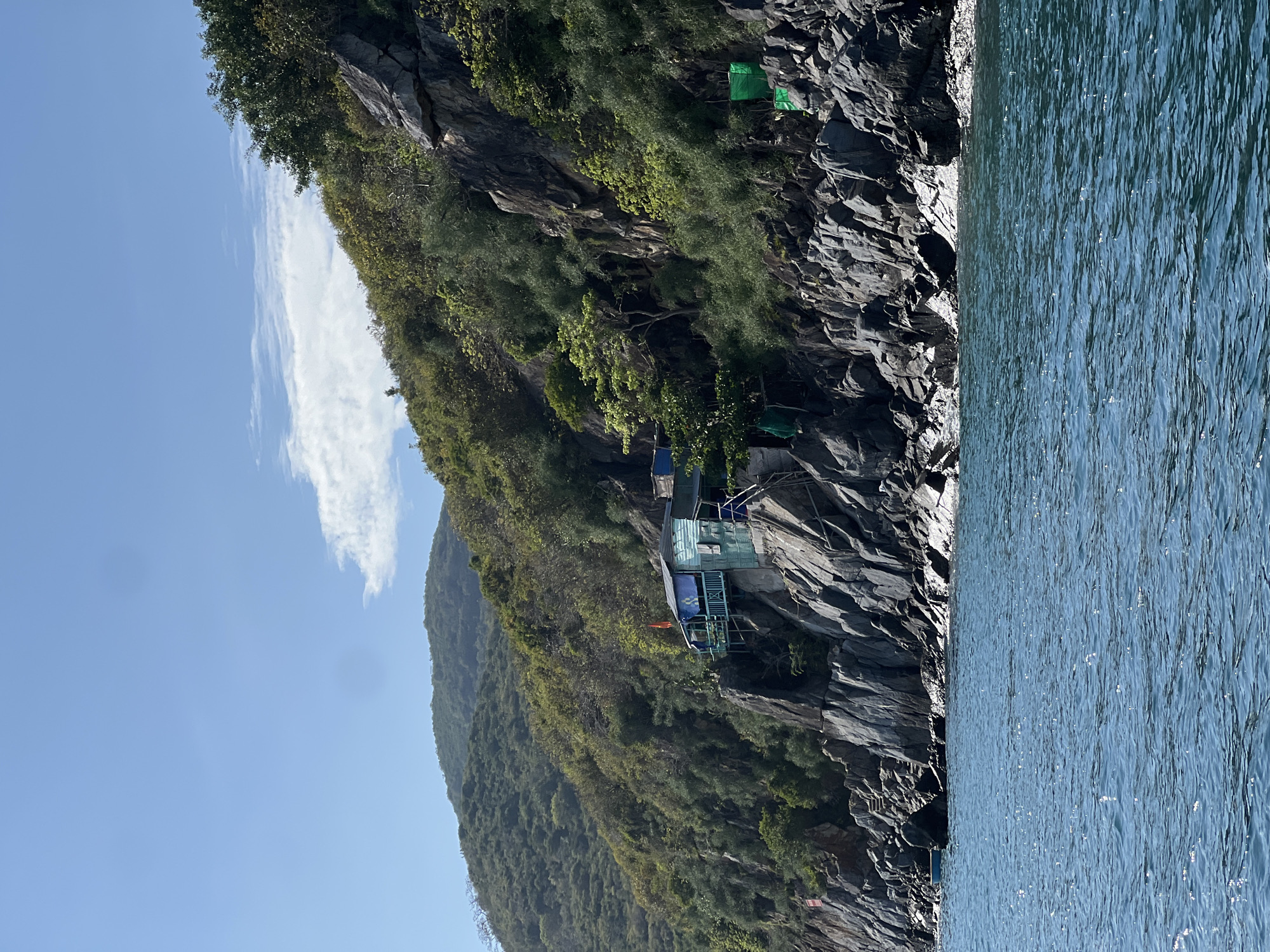 The image captures a rugged, rocky shoreline with a small, pale blue structure perched precariously on the cliffside. The structure, possibly a dwelling or a lookout point, has a visible balcony and staircase. The steep slope of the cliff is covered with dense green vegetation. The turquoise water laps against the base of the rocks, reflecting sunlight. A thin, wispy cloud stretches across the otherwise clear blue sky. The scene is located in the city of Bac Nha Trang Ward, Vietnam.