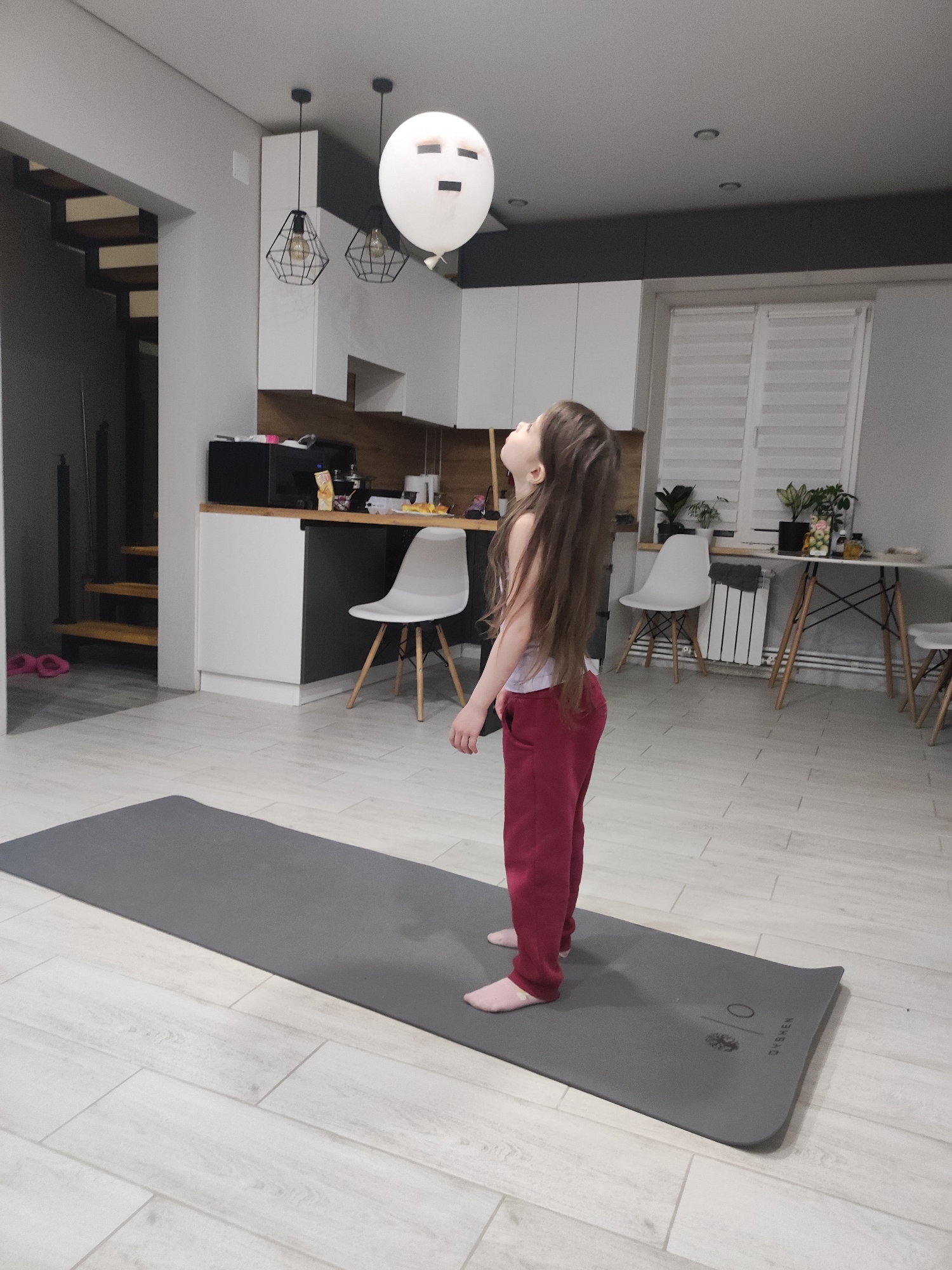 A young girl with long brown hair stands on a gray yoga mat in a brightly lit room and looks up at a white balloon with a drawn-on face. She is wearing a light-colored tank top and maroon sweatpants. The room appears to be the interior of a house or apartment, with a kitchen visible in the background, including white cabinets, a microwave, a counter with items, and white chairs. There is a dining table with plants near a window with blinds. A staircase is partially visible in the background on the left. Given the prompt's location constraint, the image shows the interior of a home in Votkinsk, Russia.