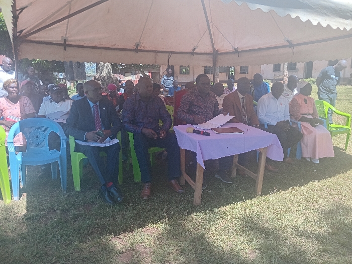 An outdoor gathering is depicted in Idakho South ward, Kenya, taking place on a grassy field under a light-colored canvas tent providing shade. In the foreground, a front row of seven individuals, comprising six men and one woman, are seated on plastic chairs (light blue and green) and other dark-colored chairs. A small rectangular wooden table draped with a light purple tablecloth is positioned in front of four of these individuals. On the table, several documents, a black microphone, and a dark-colored object are visible. A man in a dark patterned shirt is seated behind the table, looking down at papers. To his left, another man in a dark suit, light blue shirt, and patterned tie is seated, holding what appears to be a document or phone. The woman on the far right of the front row wears a light peach/pink dress and a white head covering. Behind the front row, an estimated 15-20 additional individuals, consisting of both men and women, are seated or standing under the tent, observing the front. In the background, beyond the tent structure, multiple single-story and two-story buildings, along with trees and foliage, are visible under a bright sky.