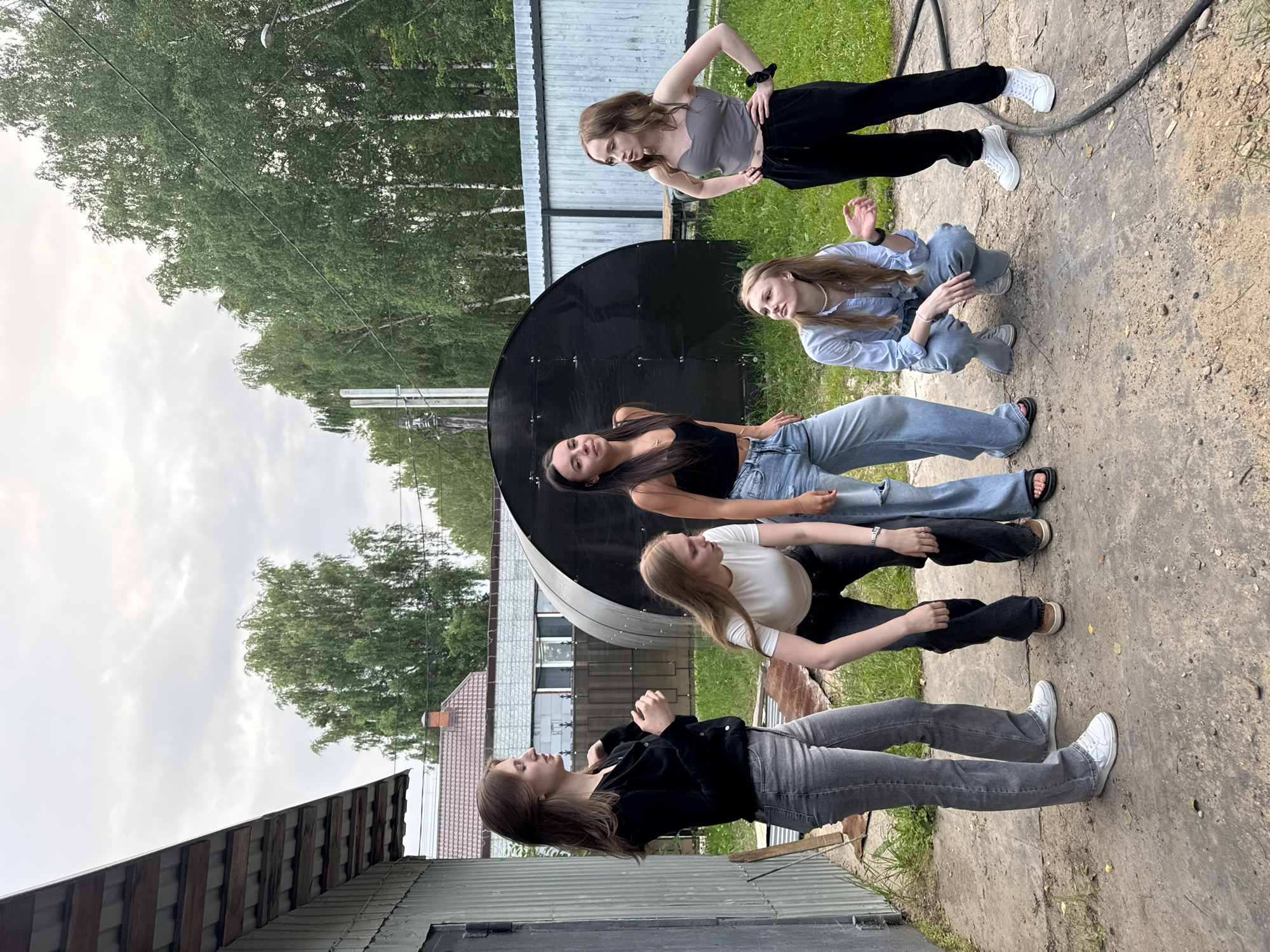 Five young women are posed outdoors on a cloudy day, likely in a rural or suburban setting in Perm, Russia. The scene is dominated by a large, black, curved object, possibly a satellite dish or a piece of equipment. The ground is a mix of dirt and concrete.
The women are dressed casually. One woman at the top of the frame wears black pants and a grey top, with her hand on her hip. Next to her, a woman in a light blue shirt and jeans is crouching. Below them, a woman in a black tank top and jeans is lying down, looking up. Another woman in a white t-shirt and black pants is also lying down, with her head turned away. The fifth woman, at the bottom of the frame, wears a black jacket and grey jeans, and is in a sitting or kneeling position.
The lighting suggests it is daytime, but the overcast sky indicates a cloudy or possibly rainy day. The background includes greenery, trees, and some simple buildings with corrugated roofs, contributing to the impression of a somewhat rustic location. There are no visible text elements in the image. The overall mood appears relaxed and informal.