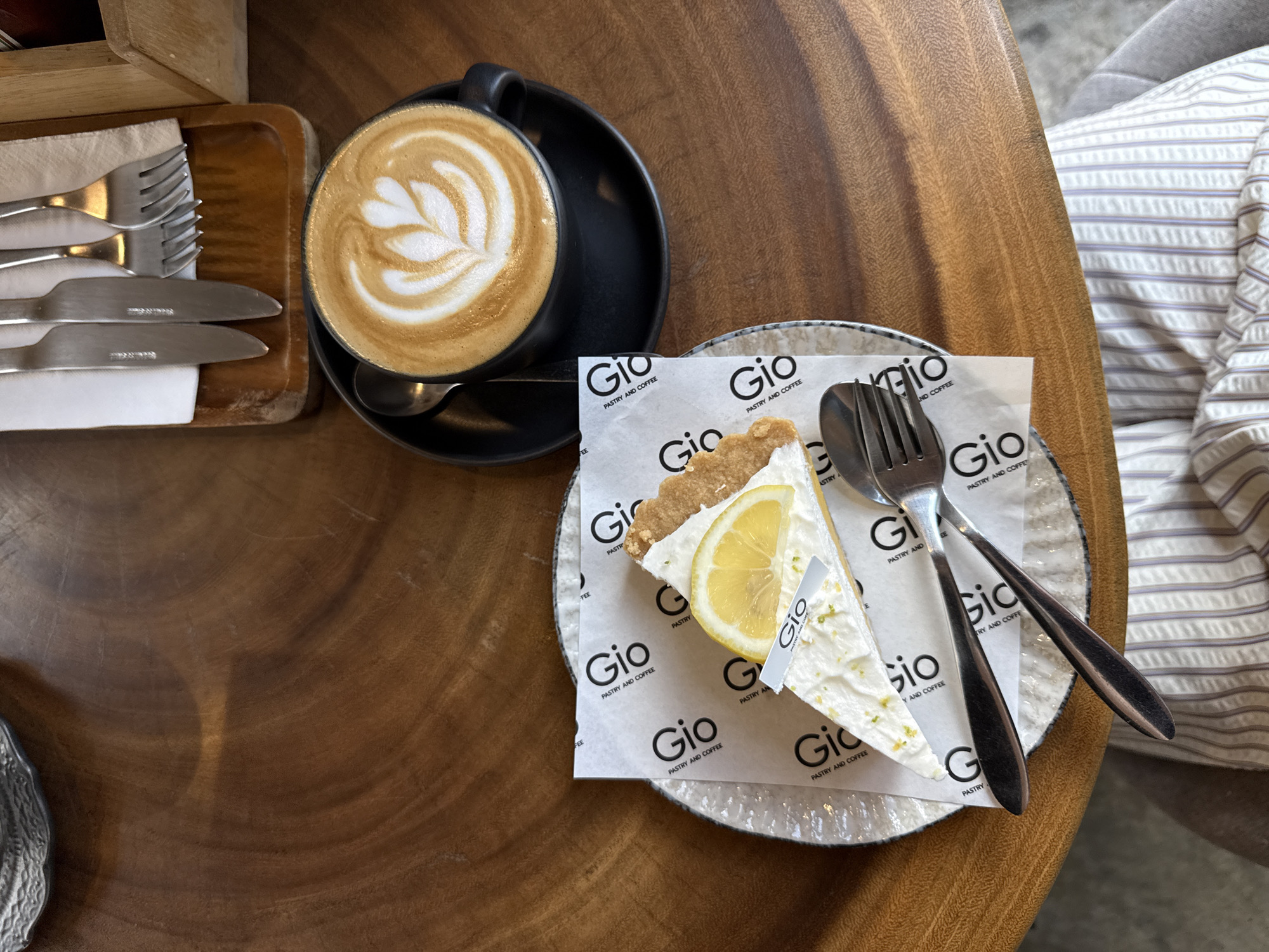A slice of lemon cheesecake topped with a lemon slice and a decorative paper flag with the text "Gio Pastry and Coffee" sits on a white paper liner on a white patterned plate. To the right of the plate is a black mug of coffee with latte art, resting on a black saucer. A spoon is visible to the left of the mug. In the background, a wooden tray holds a set of silverware, including a knife and two forks. The objects are placed on a round, polished wooden table. A person's striped clothing is visible at the bottom of the frame. The scene appears to be at a cafe in Baan Bo Phut, Thailand.