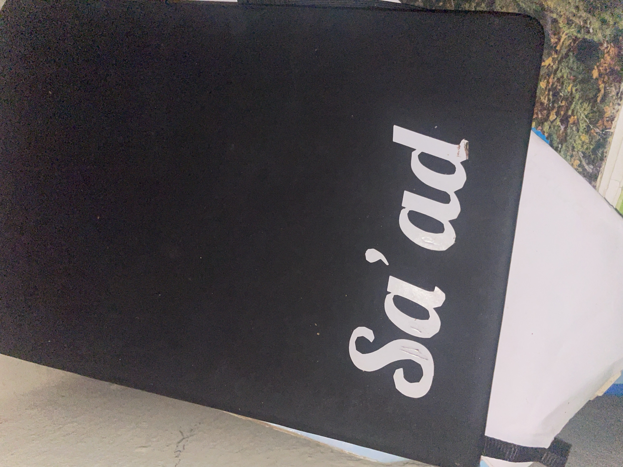 The image shows a black laptop with white text that reads "Sa'ad" on its lid. The laptop is closed and positioned at an angle. The background is blurred, but appears to be a mix of natural elements, possibly foliage, and a white surface. The scene is indoors or on a surface that is not directly exposed to the elements. There are no people visible in the image. The lighting suggests it is daytime, with the light source coming from the side, creating shadows on the laptop lid. The overall scene is casual and appears to be a close-up of an object. The location in Safari, Nigeria, is not discernible from the visual information provided in the image.