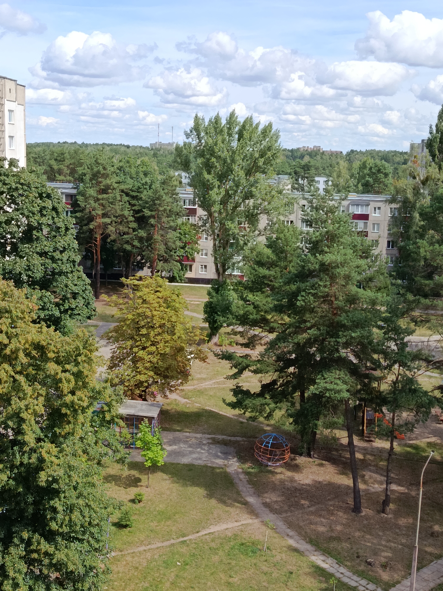 The image depicts an aerial view of a residential area in Svietlahorsk, Belarus, on a sunny day with scattered clouds. The scene is dominated by a mix of residential buildings, mature trees, and a playground. In the foreground, on the left, a large deciduous tree with vibrant green leaves partially obscures the view. Behind it, a multi-story apartment building with a light-colored facade and white-framed windows is visible.
The middle ground features a cluster of tall evergreen pine trees with dense foliage, interspersed with smaller deciduous trees displaying shades of green and yellow. These trees surround more apartment buildings, which appear to be constructed with grey concrete panels and feature red accents around some windows.
A paved pathway winds through the grass-covered ground. A small, open-sided shelter with a grey roof is situated in the middle left of the scene, with a young green tree planted nearby. Further into the middle ground, a metal spherical play structure, possibly a climbing frame, is present. To its right, a section of a playground with orange slides and red structures is partially visible.
In the background, a dense line of trees stretches across the horizon, with more apartment buildings peeking through the foliage. In the far distance, faint outlines of more buildings and possibly industrial structures can be discerned against the sky. A single lamppost with a curved top stands on the right side of the image. The overall impression is one of a well-established, green urban environment.