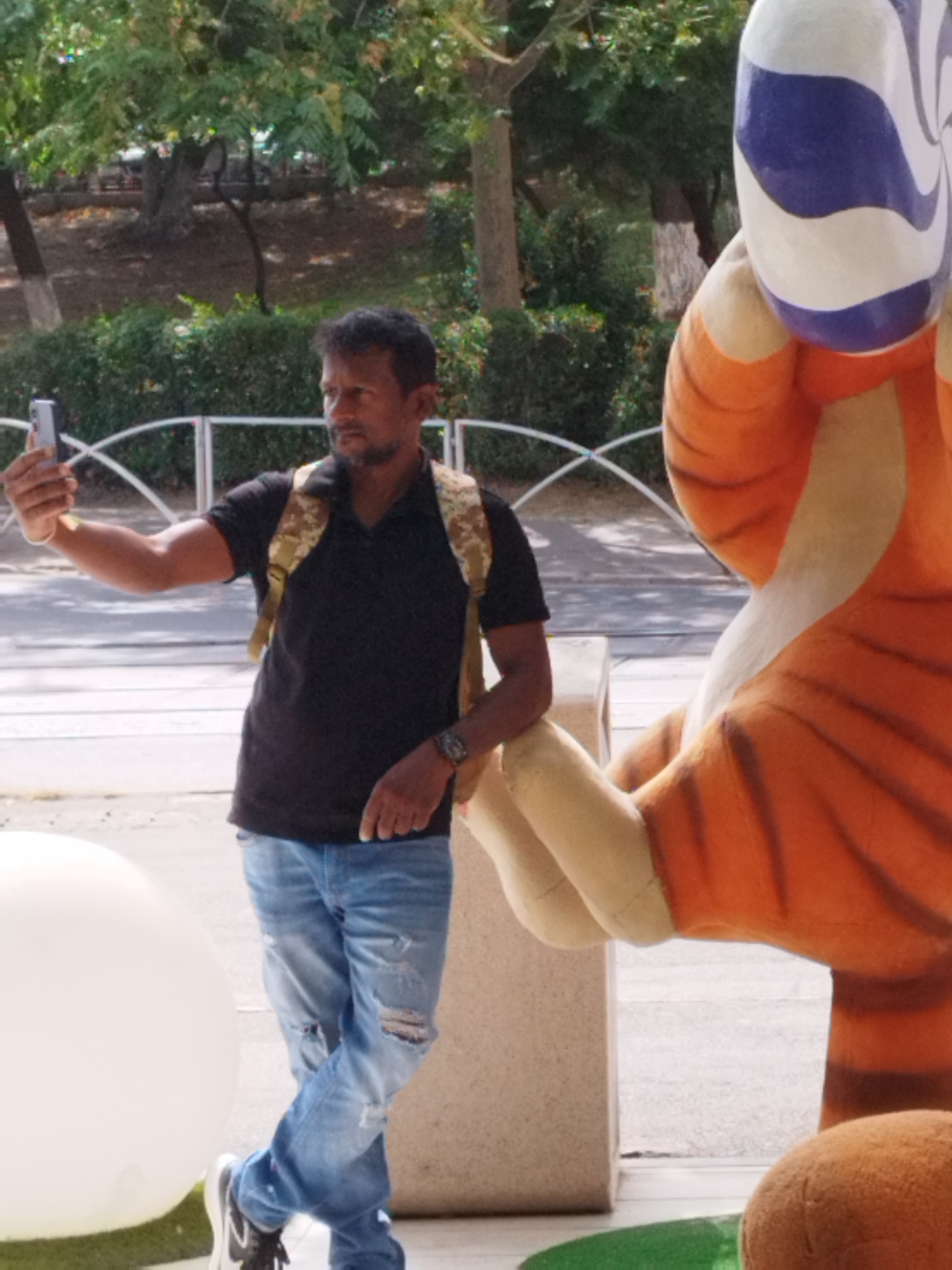 A man stands outdoors, taking a selfie with his phone. He is wearing a black polo shirt, ripped blue jeans, a camouflage backpack, and a watch on his left wrist. To his right is a large, cartoonish sculpture of a tiger, with orange and white stripes. In the background, there are trees, bushes, a white fence, and a building. The scene is lit by natural daylight. The location is confirmed to be Bucharest, Romania.