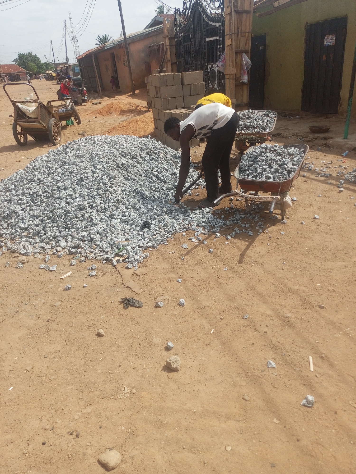 The image shows a construction site in Yelwa, Nigeria. A man wearing a white shirt and black pants is filling a wheelbarrow with crushed stone from a large pile on a sandy ground. Another wheelbarrow, already filled with crushed stone, is nearby. In the background, there are simple buildings, a stack of concrete blocks, a metal gate, and a wooden cart. Power lines are visible overhead. A second person is wearing a yellow shirt.