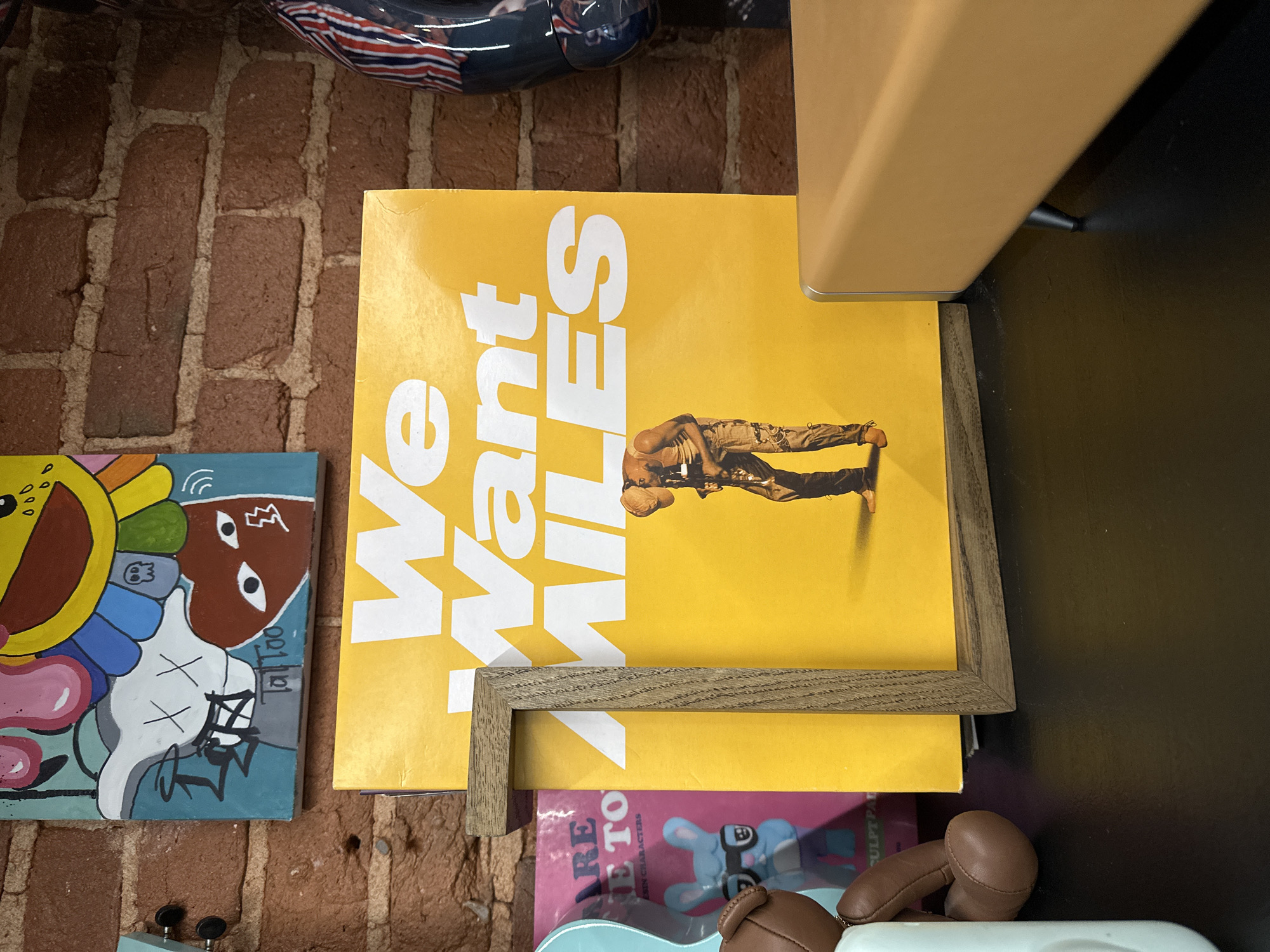 The image displays a still life arrangement of art and design objects, potentially on a shelf or table with a brick wall backdrop. A yellow vinyl record cover prominently features the text "We Want MILES" and an image of a man. The record is partially framed by a light-colored wooden frame. To the left, a colorful painting with cartoonish characters reminiscent of KAWS's style is visible. Below the record, glimpses of another artwork or book with pink and blue hues are visible. A section of a leather-like object (possibly a toy or decorative piece) is also present. The setting does not appear to be in Moscow, Russia, as there are no readily identifiable signs or markers indicative of Moscow's specific urban environment, landmarks, or architecture. The details lean toward an interior decorative space rather than a discernible public or commercial place in Moscow.
