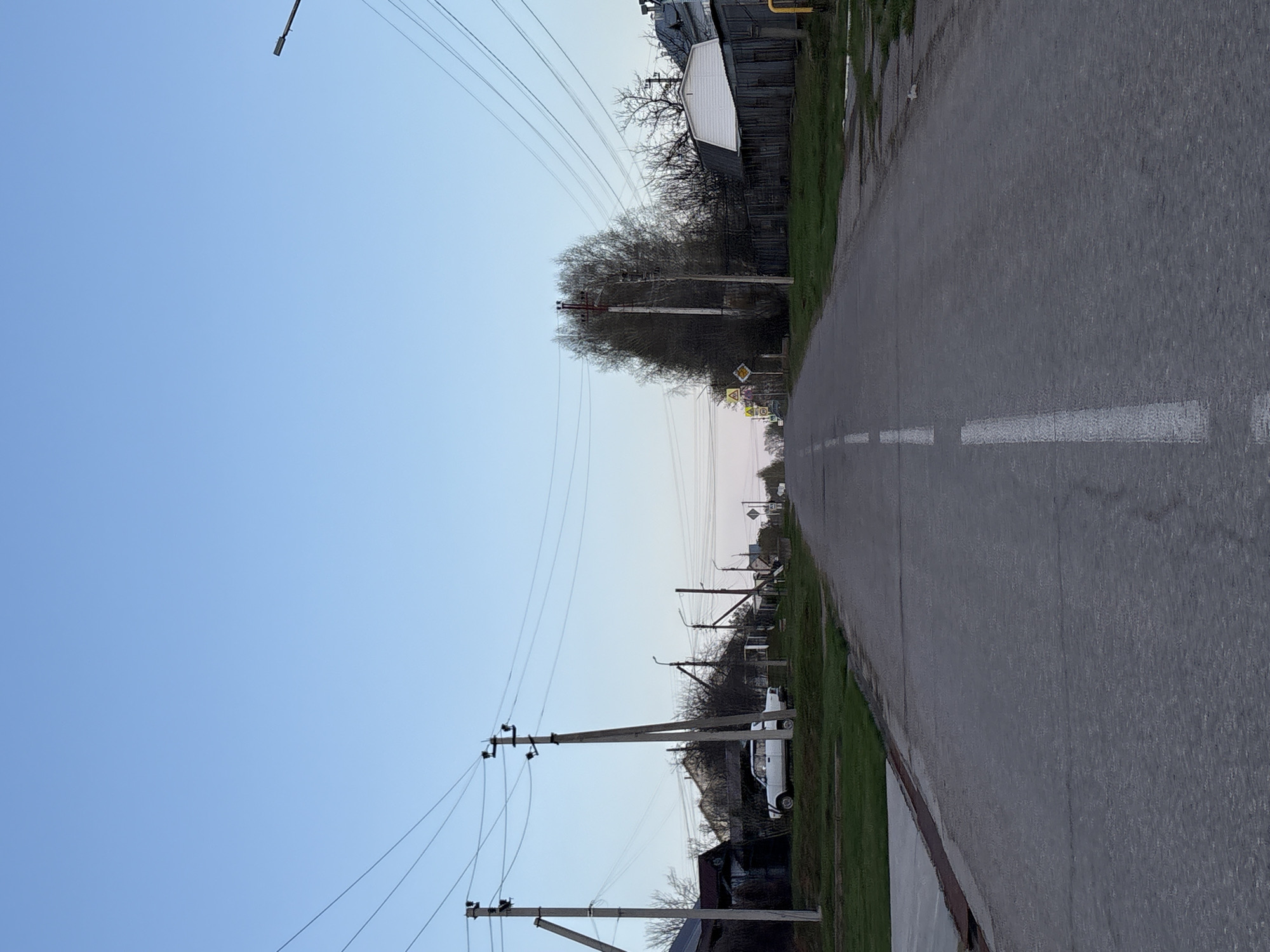 An outdoor shot captures a street in Прикамский, Russia. The street is asphalt, with a white broken centerline. Power lines run above the street, supported by utility poles. Buildings and vegetation line the side of the road. The sky is light blue.