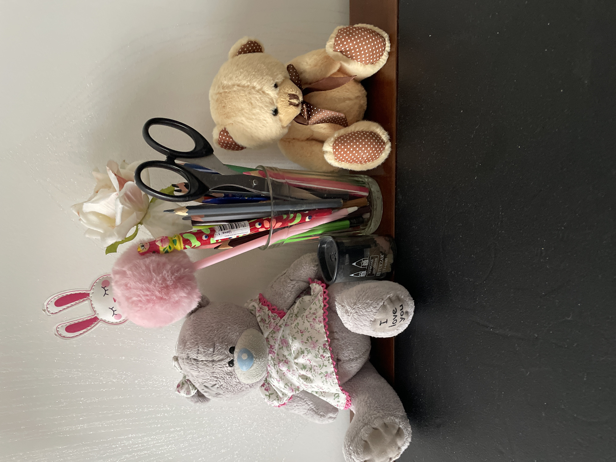 The image presents a tabletop scene, likely indoors. On the left side of the image is a white surface. A gray teddy bear wearing a floral-patterned dress and a pink rabbit-shaped pom-pom are placed here. Also present is a glass container filled with pencils and a pair of scissors. Resting near this are some white flowers. On the right side of the image, there is a dark, possibly black, surface. On the top of the image, there is a tan teddy bear with brown polka-dot paws and a brown bow. The photo seems to have been taken in Лесной Пейзаж Кп (шарапово, Russia.