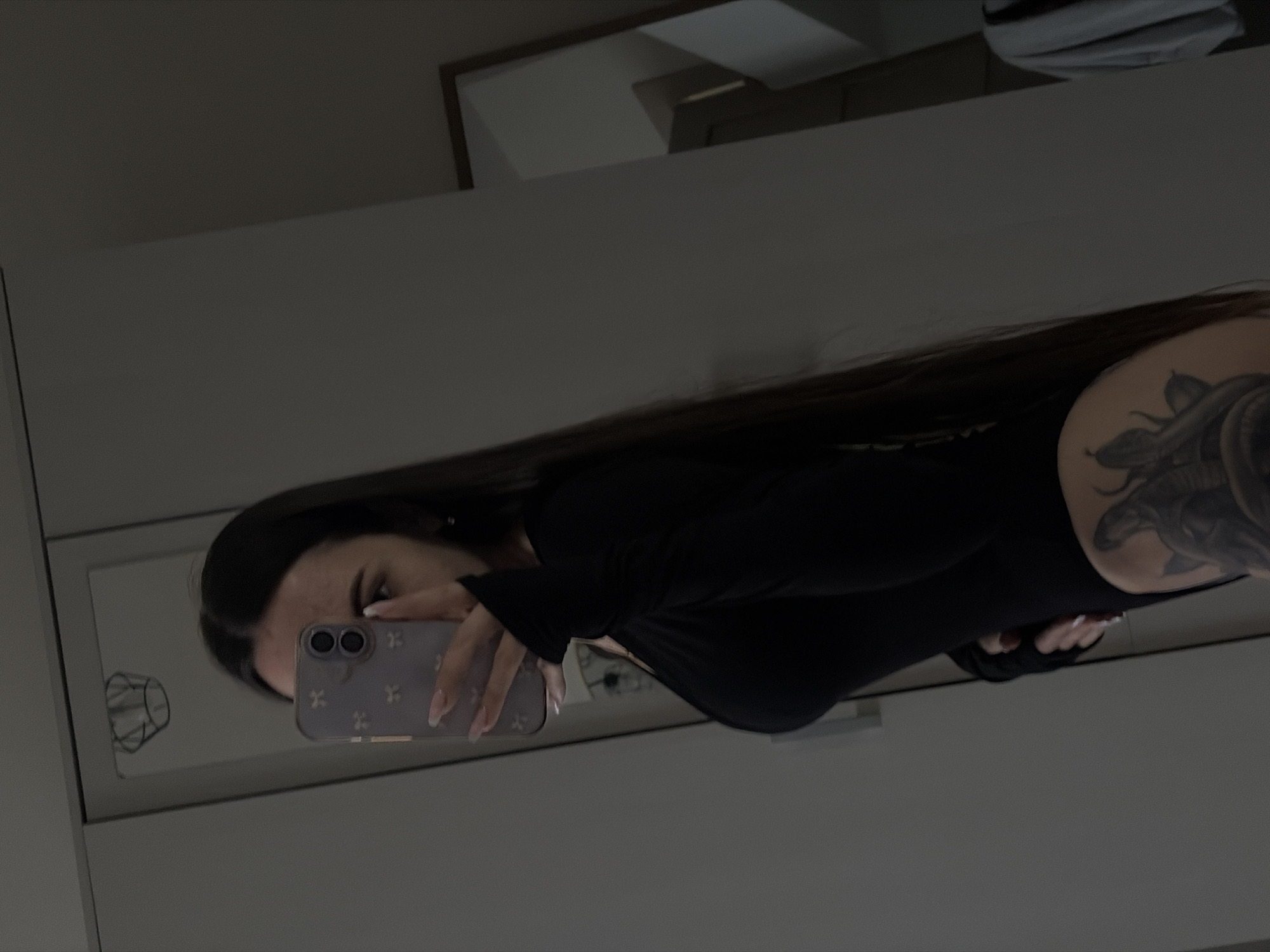 The image, presented with a landscape orientation (after correcting the original rotation), shows a young woman taking a mirror selfie indoors. Her face, featuring her eyes and nose, is partially visible in the reflection of a rectangular mirror. She has long, dark hair and is wearing a form-fitting, long-sleeved black top or bodysuit. A prominent, detailed tattoo, possibly depicting a snake or similar creature, covers her right shoulder and upper arm.
She is holding a light purple smartphone, equipped with a multi-lens camera system and a case adorned with a pattern of small 'X' or cross-like symbols, in her left hand to capture her reflection. Her fingernails are long and manicured with a light, neutral polish. The mirror also reflects a simple line drawing, resembling a geometric object, in its lower-left corner.
The setting is an interior room, likely a home, with plain, light grey walls or furniture forming the background. In the upper right background, a dark frame is partially visible, reflecting a lighter area. The lighting is relatively dim, creating a subdued ambiance. While no specific architectural or environmental cues within the image confirm the location, the provided context indicates the scene is set in Saint Petersburg, Russia. No legible text is visible in the image.
