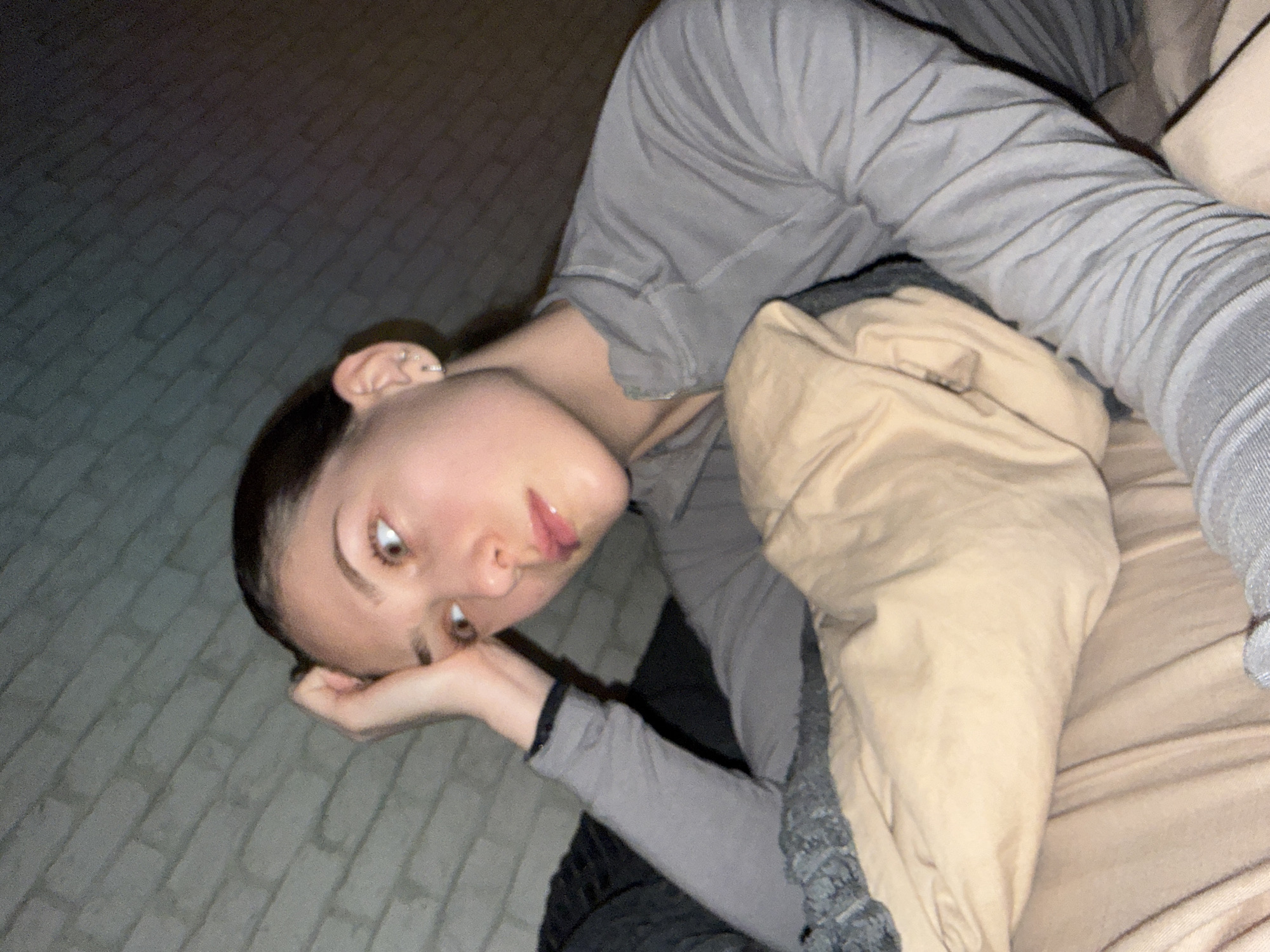 A young woman lies on a bed or sofa covered in beige and gray bedding. She wears a long-sleeved gray shirt or pajama top. Her head is tilted towards the left side of the frame, and her left hand rests on a paved surface resembling a sidewalk or patio. The backdrop suggests an outdoor setting, possibly a balcony or terrace. The image is taken at an angle, with a slight rotation. There is no specific indication to determine if it is taken in the city of Sofia, Bulgaria.