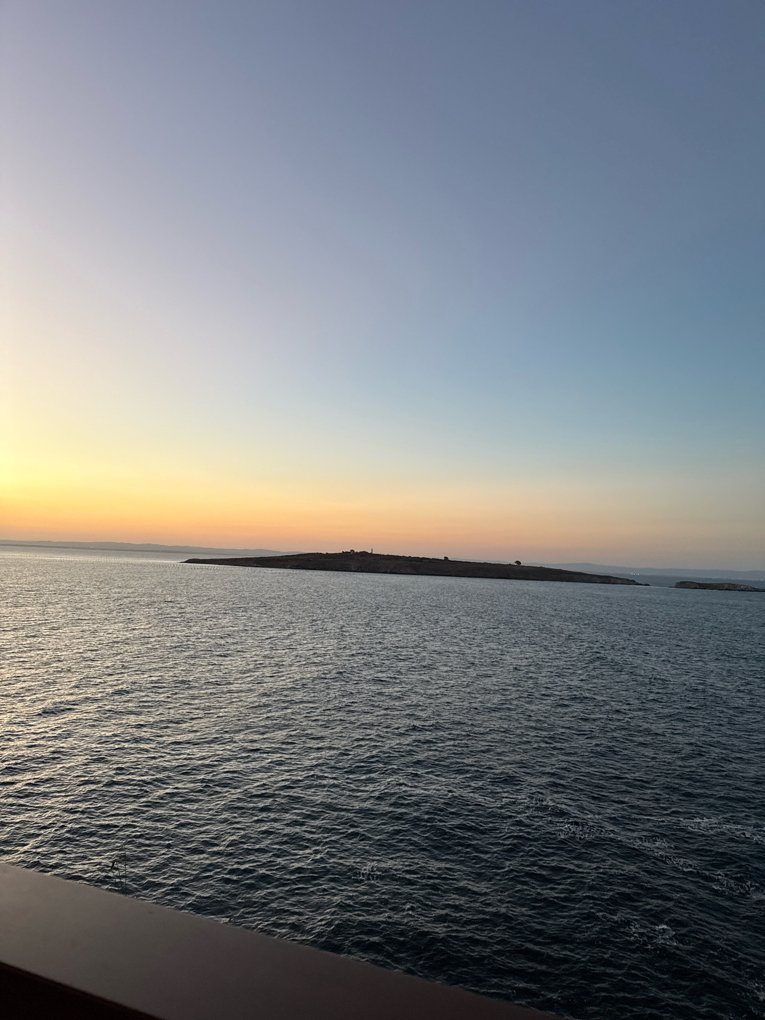 A wide shot of the sea off the coast of Sozopol, Bulgaria, captures a serene sunset. The sky transitions from a pale yellow and orange near the horizon to a soft blue overhead. A long, thin island is visible in the distance, dotted with sparse vegetation and a small structure, possibly a lighthouse, near its center. The water is a deep blue, rippling gently. In the foreground, the edge of a dark, horizontal surface, likely part of a boat or balcony, is visible.
