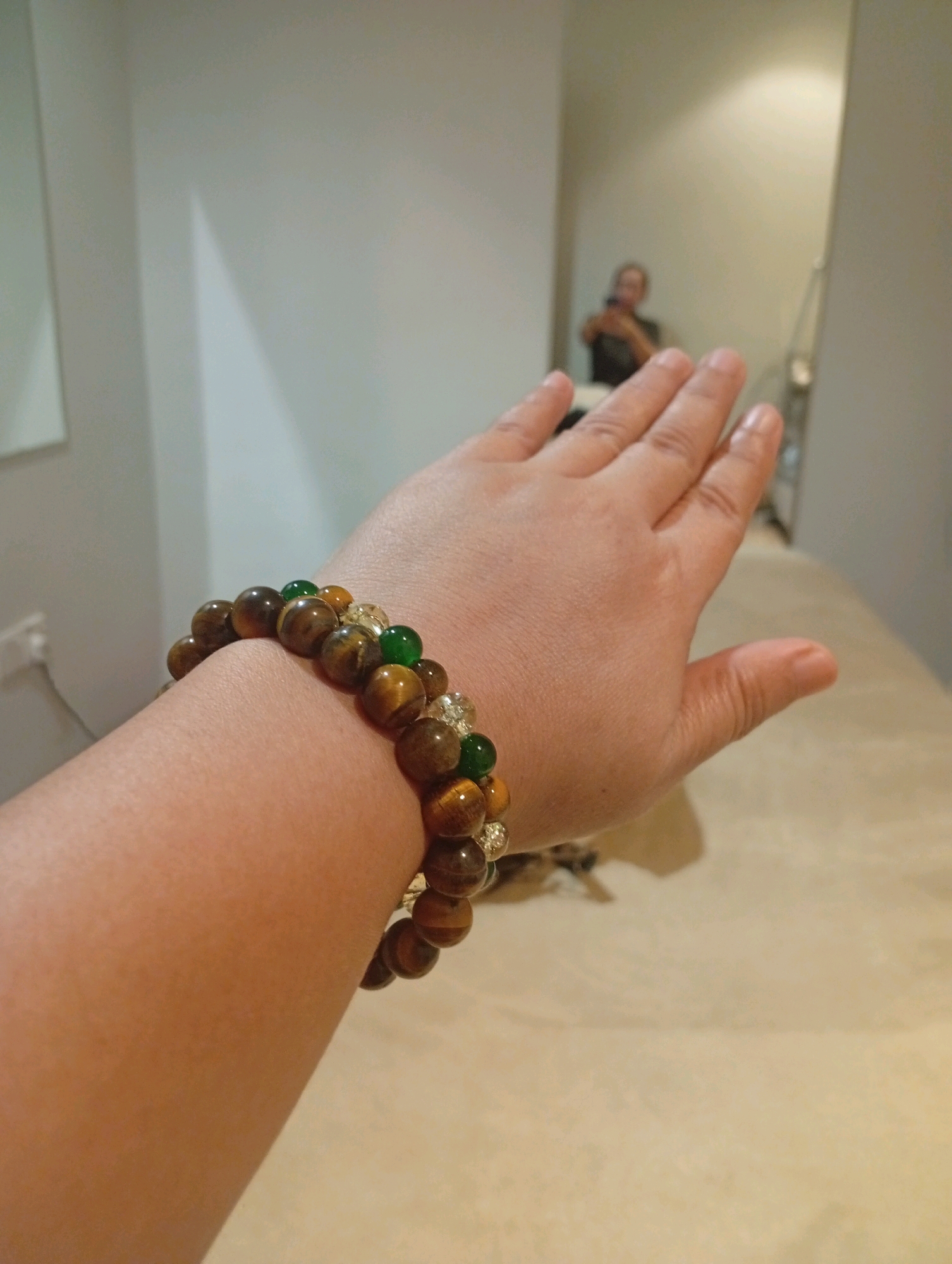 The image shows a person's left arm with a bracelet made of brown and green beads, along with some clear beads. The arm is extended towards the camera. In the background, a blurred reflection of another person holding a phone can be seen in a mirror. The environment appears to be an indoor space, possibly a room with light-colored walls and a textured surface in the foreground. The location in Selayang Municipal Council, Malaysia, cannot be determined from the visual information provided in the image.