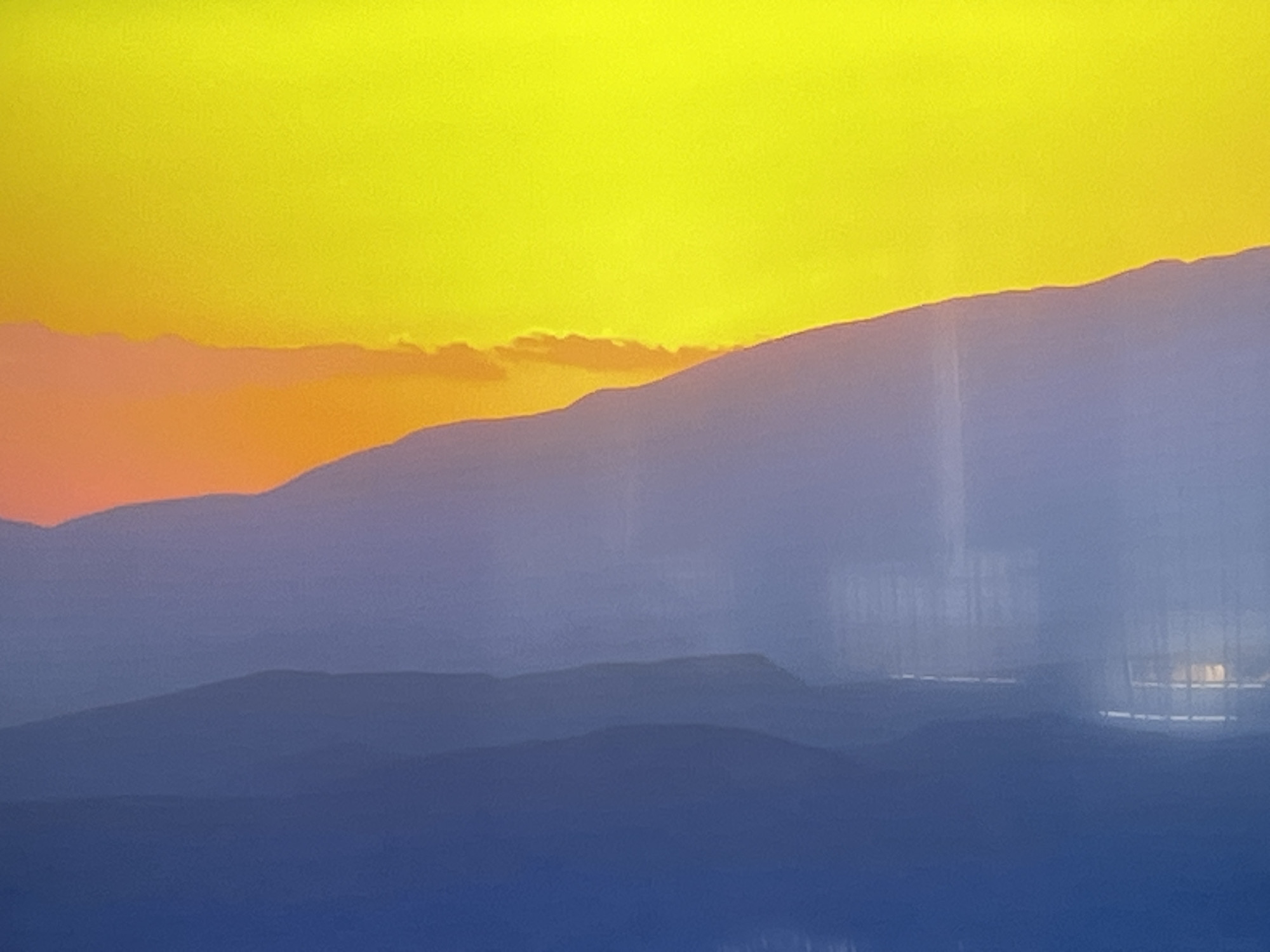 The image depicts a layered landscape of mountains or hills against a bright yellow and orange sky, suggesting a sunset or sunrise. The foreground is a gradient of blue, transitioning from a deep blue at the bottom to lighter blues and purples in the mid-ground. These layers of hills are silhouetted and indistinct due to atmospheric haze. In the upper portion of the image, the sky is a vibrant yellow, with a band of orange clouds or haze visible at the horizon line where it meets the highest mountain range. There are no visible people, subjects, or objects that indicate a specific location within the city of Kharkiv, Ukraine. The image is an abstract landscape photograph.