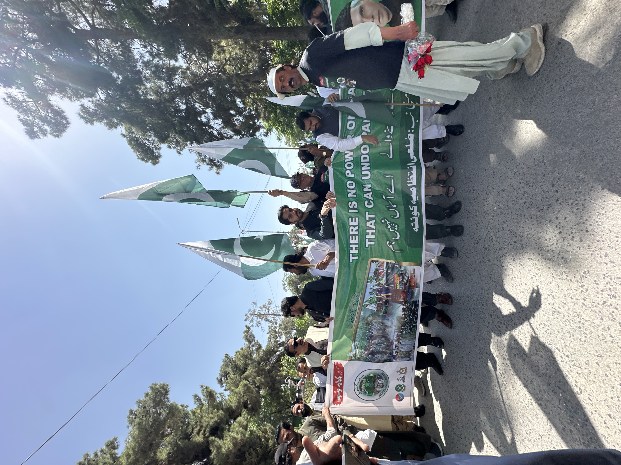 A group of men are marching in Quetta City Tehsil, Pakistan. They are carrying Pakistani flags and a banner with the text "THERE IS NO POWER ON THAT CAN UNDO PAKISTAN". One man holds a picture of a woman and red flowers. The weather is sunny, and there are trees in the background.