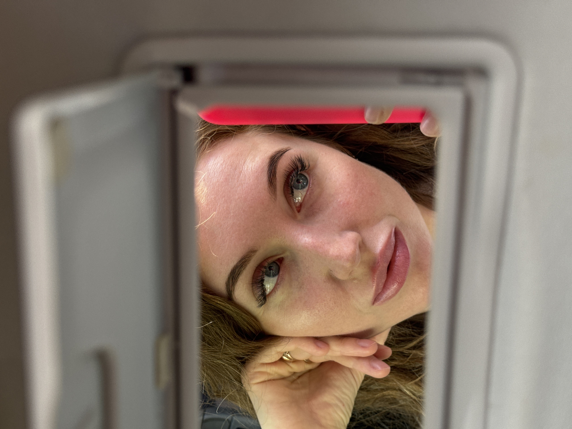 A woman with fair skin and light brown hair is looking at the camera through the open mirror of a car visor. Her face is partially framed by the open visor door, which is gray. She has long eyelashes and is wearing lipstick. A hand with a gold ring is visible near her cheek. A pink object is visible behind the top edge of the visor. It is not possible to determine the location in Chaikovsky, Russia.