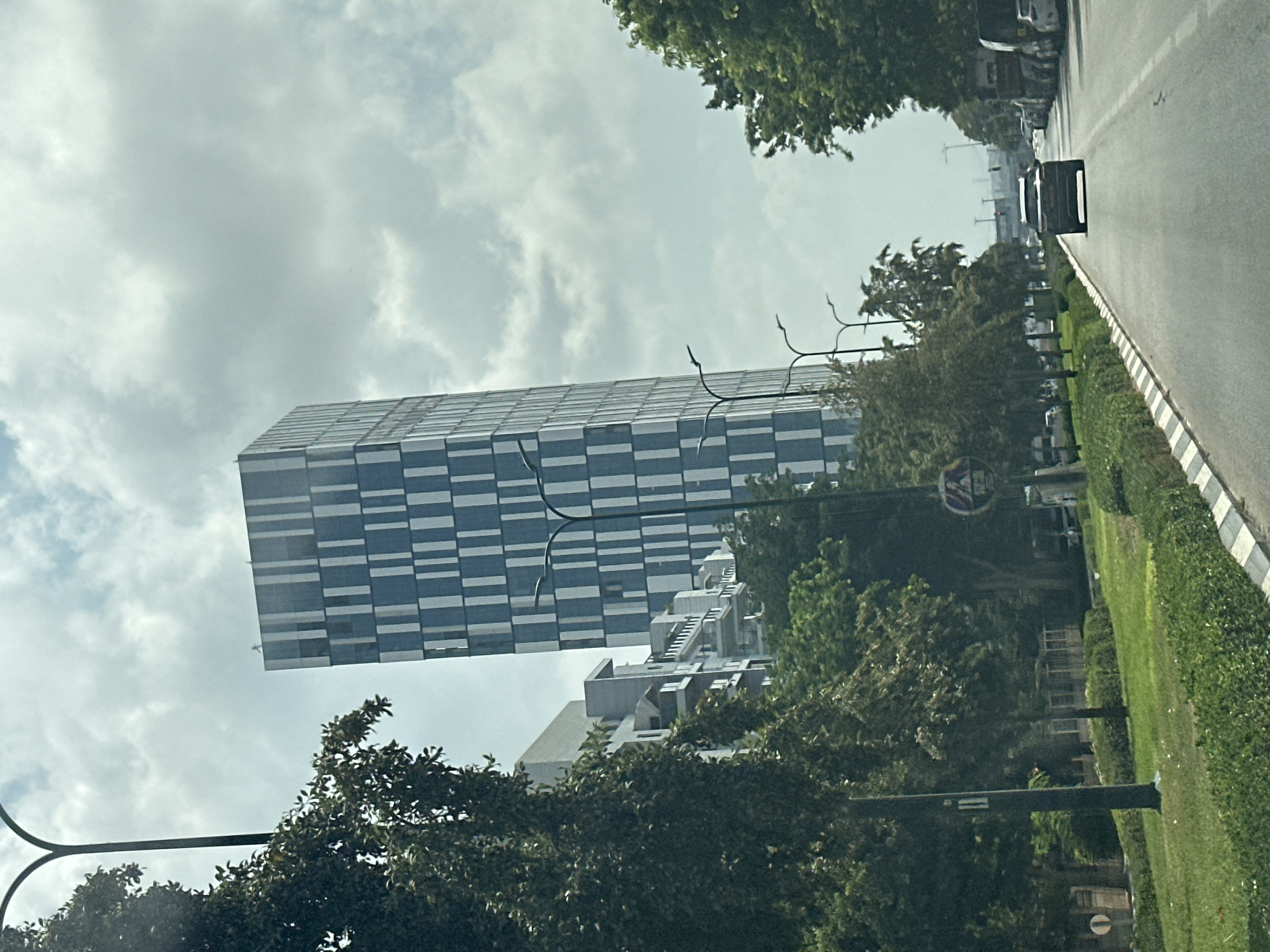 The image depicts a street scene in Netanya, Israel, likely on a cloudy day. A tall, modern skyscraper with a facade of alternating blue and white rectangular panels dominates the left side of the frame. Adjacent to it, and slightly closer to the viewer, is a multi-story building with balconies and a lighter-colored facade. A street with cars is visible in the foreground, with one dark SUV in the center lane. Trees line the street on both sides, with a grassy median separating the lanes on the left. Streetlights with curved arms are positioned along the road. In the distance, more buildings and trees can be seen, along with what appear to be traffic signals.