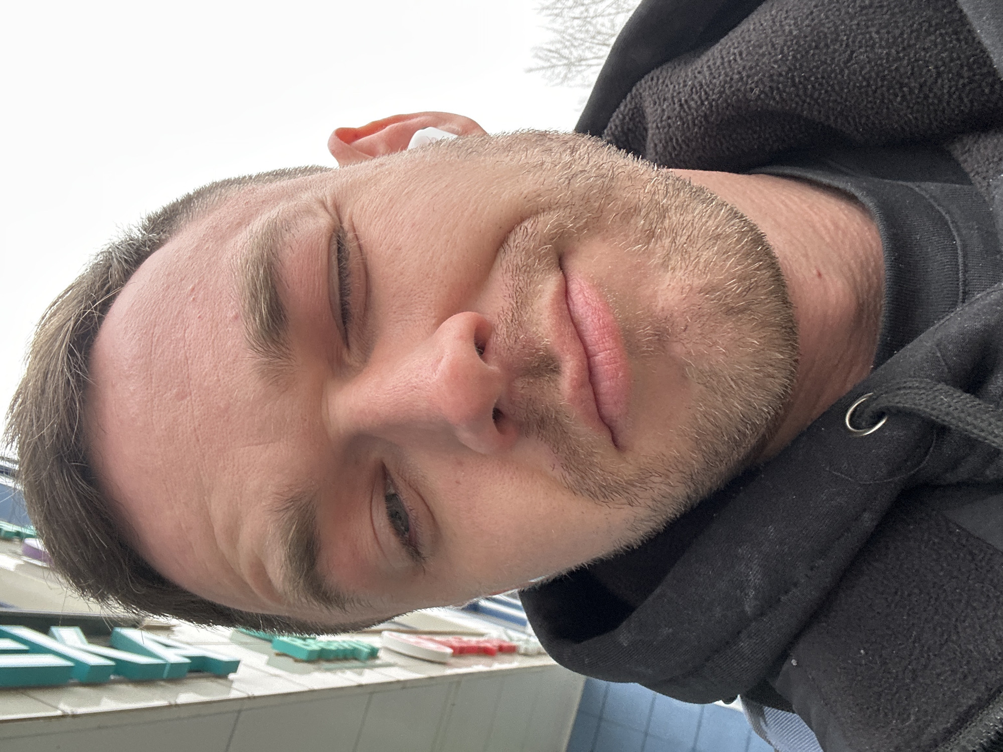 A close-up selfie of a fair-skinned man with stubble, wearing a black hooded sweatshirt, is angled upwards. He appears to be outdoors, with the sky visible in the upper part of the frame. His eyes are partially closed, suggesting a squint. In the lower part of the image, there is what appears to be a sign with teal and white lettering. This sign indicates the location is Perm, Russia, as the color scheme and lettering style of the location are similar to those used on the Perm city sign.